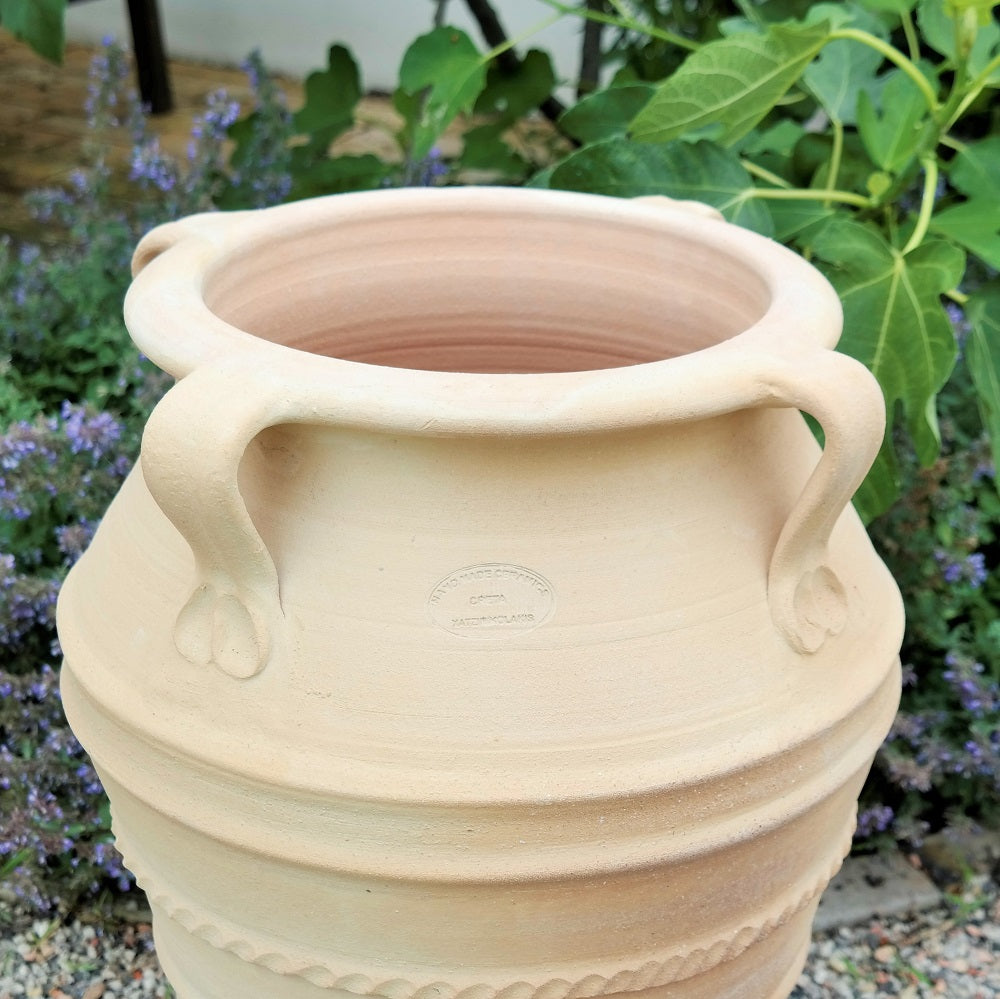 Die frostfeste Gartenamphore "Datura" (H=60 cm) in Hellbeige mit drei dekorativen Henkeln, strukturierten Bändern und geprägter Marke steht im Freien neben Grünpflanzen und lila Blüten und bringt mediterranen Charme in Ihren Garten.