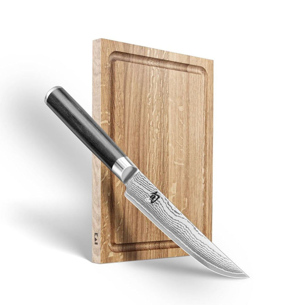 Das KAI Steakmesser+ Steakbrett "THE MAGIC KAI WISHLIST 2025" steht aufrecht hinter einem scharfen Küchenmesser mit schwarzem Griff und gemusterter Klinge vor weißem Hintergrund.
