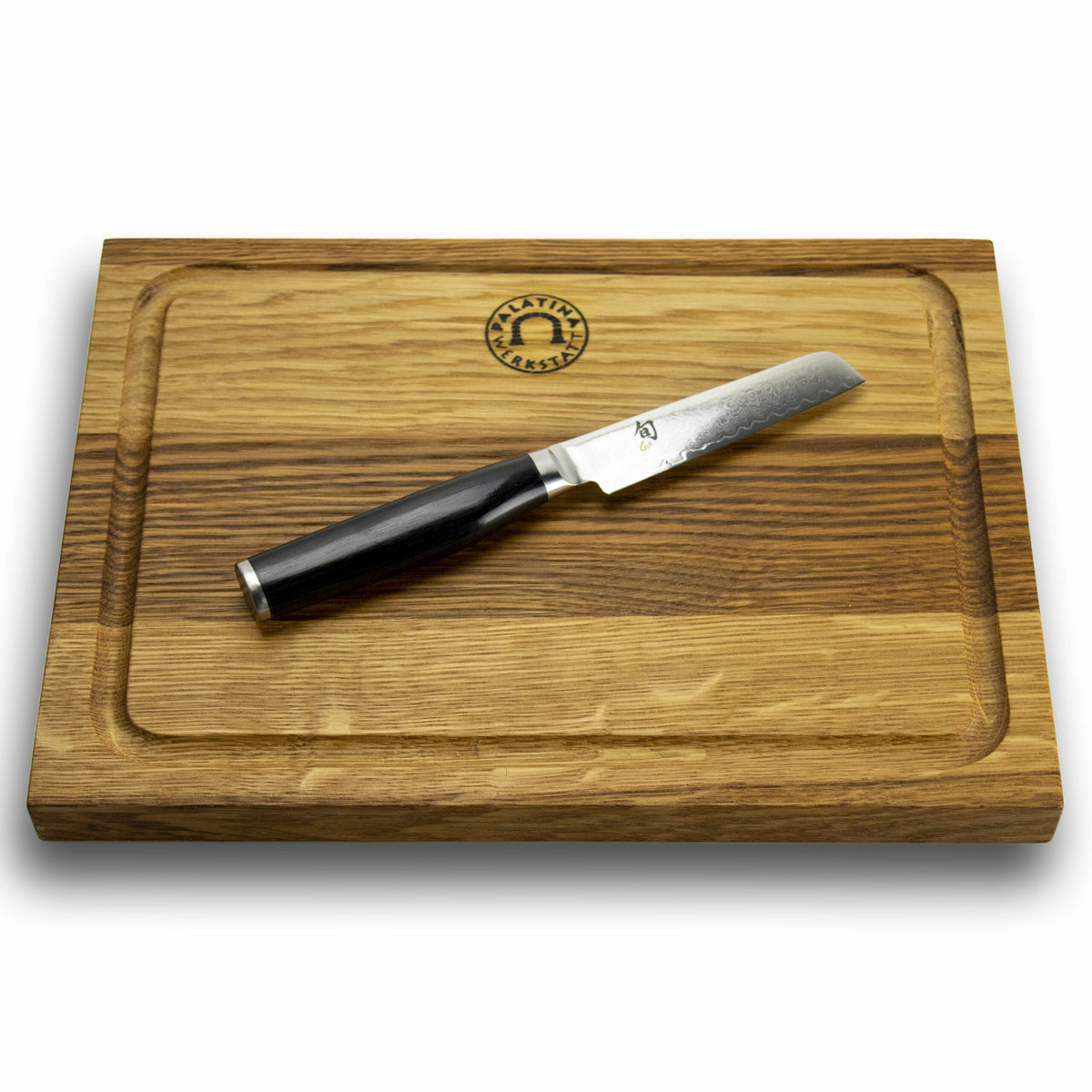 Ein Holzschneidebrett aus Eiche mit den Maßen 30x22 cm weist eine Nut um die Kante und oben in der Mitte ein Logo auf. Diagonal auf dem Brett liegt ein Kai Shun Premier Minamo TMM-0700 Officemesser, das seinen dunklen Griff und die glänzende, gemusterte 9 cm lange Damaststahlklinge zur Schau stellt.