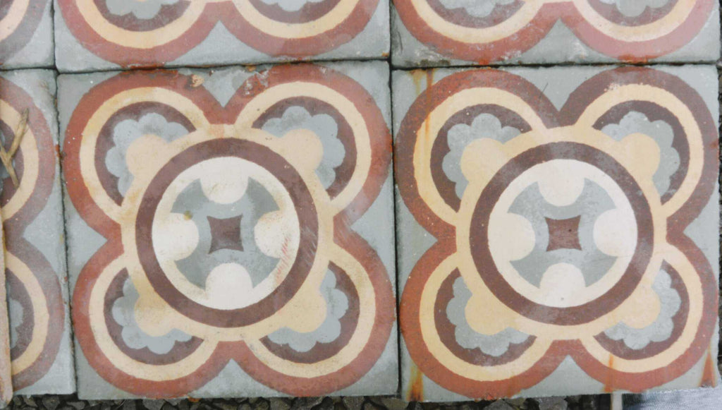 Vier Historische Gründerzeitfliesen, jeweils 16,5 x 16,5 cm groß, weisen ein symmetrisches, florales Muster in erdigen Farben wie Rot, Hellbraun, Braun und Blau auf. Jede Fliese weist eine zentrale Sternform auf, die von geschichteten Schleifen umgeben ist, wodurch ein ornamentales Design entsteht. Die leicht abgenutzten Fliesen haben sichtbare Kanten, die zu ihrem Vintage-Charme beitragen, und bedecken insgesamt etwa 17 Quadratmeter.