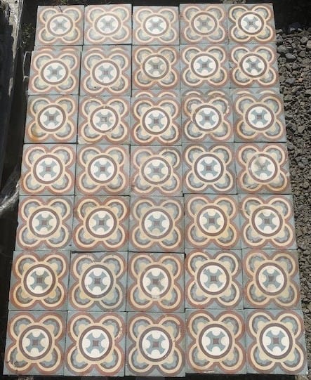 Eine rechteckige Anordnung dekorativer Fliesen mit einem sich wiederholenden Muster aus miteinander verbundenen Blumenformen. Die Historischen Gründerzeitfliesen (16,5 x 16,5 cm, ca. 5,5 m²) haben eine gedämpfte Farbpalette mit Beige-, Blau- und Orangetönen und sind in einem Raster auf einer flachen Oberfläche angeordnet. Auf der rechten Seite der Fliesen sind Kieselsteine sichtbar.