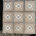 Ein Raster aus historischen Gründerzeitfliesen, wobei jede 16,5 x 16,5 cm große Fliese ein kompliziertes symmetrisches Muster in Blau-, Beige-, Creme- und Brauntönen aufweist. Inspiriert von historischen Fliesen zeigt jedes Stück ein zentrales kleeblattähnliches Motiv, umgeben von ineinandergreifenden kreisförmigen Formen vor einem strukturierten grauen Steinhintergrund. Das gesamte Paket umfasst ungefähr 5,5 Quadratmeter.