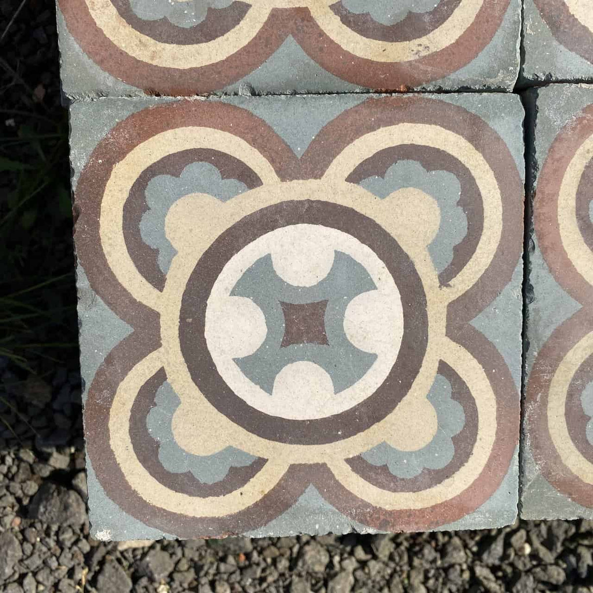Die Historischen Gründerzeitfliesen sind quadratische Keramikfliesen im Format 16,5 x 16,5 cm mit einem symmetrischen, kreisförmigen Muster mit ineinandergreifenden Formen in Blau-, Braun-, Beige- und Cremetönen. Ihr Design vereint geometrische und florale Elemente und schafft so ein komplexes und dekoratives Motiv, das von historischen Fliesen inspiriert ist. Die Fliese wird auf einer Kiesfläche präsentiert.