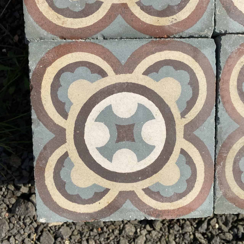 Die Historischen Gründerzeitfliesen sind quadratische Keramikfliesen im Format 16,5 x 16,5 cm mit einem symmetrischen, kreisförmigen Muster mit ineinandergreifenden Formen in Blau-, Braun-, Beige- und Cremetönen. Ihr Design vereint geometrische und florale Elemente und schafft so ein komplexes und dekoratives Motiv, das von historischen Fliesen inspiriert ist. Die Fliese wird auf einer Kiesfläche präsentiert.