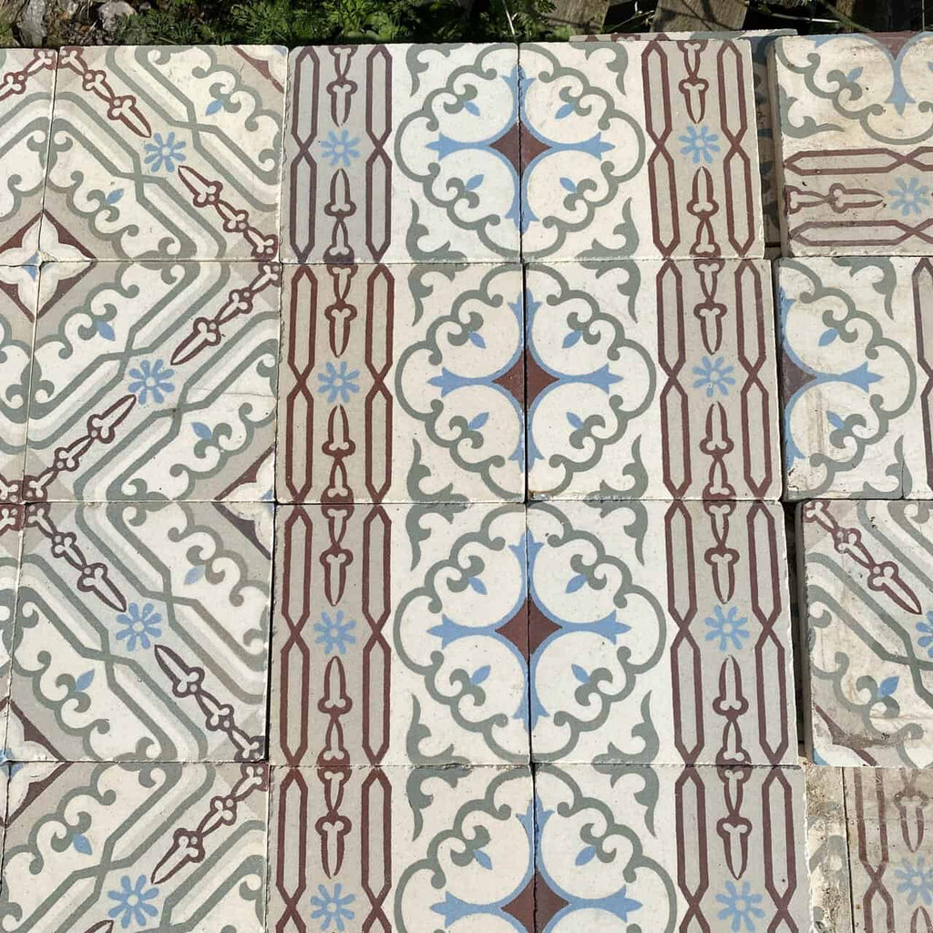 Ein Abschnitt gemusterter Bodenfliesen mit Vintage-Design. Diese *Historischen Gründerzeitfliesen* zeigen symmetrische, geometrische Muster in Blau-, Braun- und Grautönen auf hellem Hintergrund. Das zentrale Motiv umfasst Vierpassformen und ineinandergreifende Schnörkel, umgeben von linearen Akzenten. Jede Fliese misst 14 x 14 cm und deckt ungefähr 10 Quadratmeter ab.