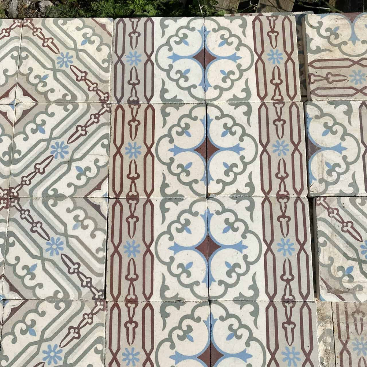 Ein Abschnitt gemusterter Bodenfliesen mit Vintage-Design. Diese *Historischen Gründerzeitfliesen* zeigen symmetrische, geometrische Muster in Blau-, Braun- und Grautönen auf hellem Hintergrund. Das zentrale Motiv umfasst Vierpassformen und ineinandergreifende Schnörkel, umgeben von linearen Akzenten. Jede Fliese misst 14 x 14 cm und deckt ungefähr 10 Quadratmeter ab.