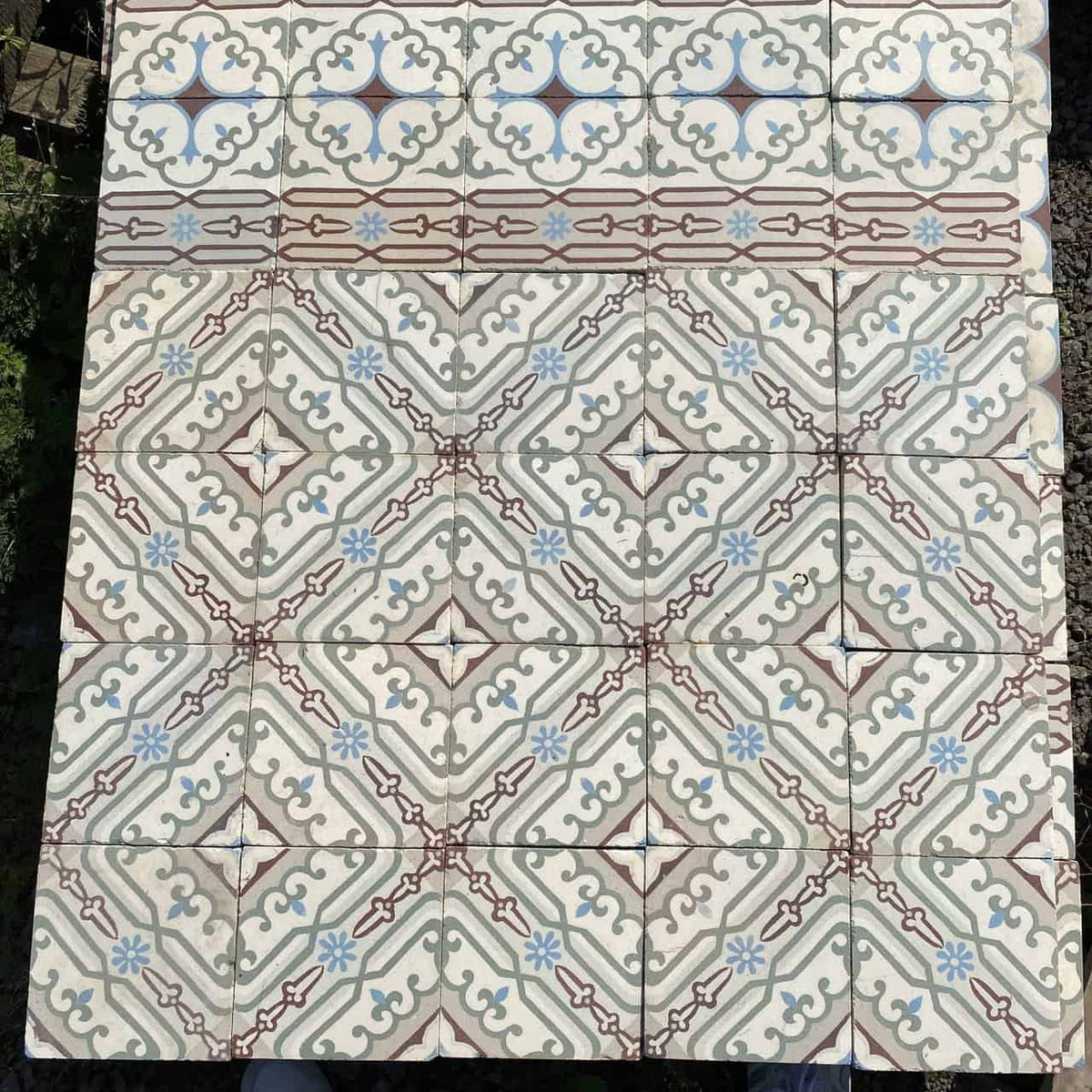 Eine Detailansicht quadratischer Bodenfliesen mit der Marke Historische Gründerzeitfliesen, die ein kompliziertes geometrisches Mosaikmuster zeigen. Das Design umfasst symmetrische Formen in Beige-, Blau- und Kastanienbrauntönen, die an historische Fliesen erinnern und ein sich wiederholendes Rauten- und Kreuzmotiv bilden. Die oberste Reihe weist einen dekorativen Rand in Blau und Kastanienbraun auf. Jede Fliese misst 14 x 14 cm und deckt ungefähr 10 Quadratmeter ab.