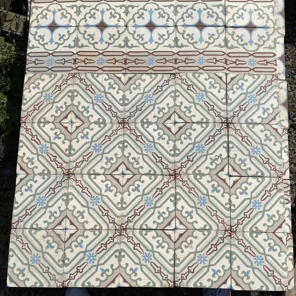 Eine Detailansicht quadratischer Bodenfliesen mit der Marke Historische Gründerzeitfliesen, die ein kompliziertes geometrisches Mosaikmuster zeigen. Das Design umfasst symmetrische Formen in Beige-, Blau- und Kastanienbrauntönen, die an historische Fliesen erinnern und ein sich wiederholendes Rauten- und Kreuzmotiv bilden. Die oberste Reihe weist einen dekorativen Rand in Blau und Kastanienbraun auf. Jede Fliese misst 14 x 14 cm und deckt ungefähr 10 Quadratmeter ab.