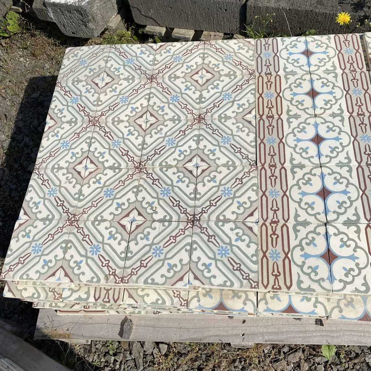 Ein Stapel dekorativer historischer Gründerzeitfliesen, 14 x 14 cm groß und ungefähr 10 Quadratmeter groß, mit komplizierten geometrischen Mustern in Blau, Weiß, Braun und Hellgrün. Die Designs umfassen Rauten, Kreuze und Blumenmotive. Die Fliesen liegen auf einer Holzpalette auf einem Kiesboden, daneben steht eine einzelne gelbe Blume.