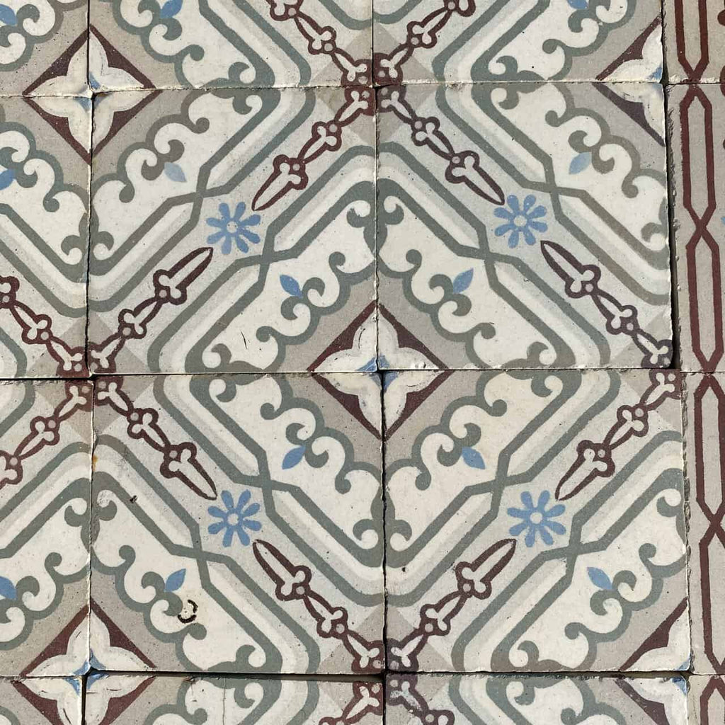 Eine Nahaufnahme von Historischen Gründerzeitfliesen mit blassblauen Blumenmustern und kastanienbraunen und grauen geometrischen Formen. Die Fliesen, die jeweils 14 x 14 cm groß sind und etwa 10 Quadratmeter bedecken, bilden ein sich wiederholendes symmetrisches Muster und erzeugen einen Vintage-Dekorationslook, der an historische Fliesen erinnert. Die Fugenlinien sind leicht ungleichmäßig, was den rustikalen Charme der Fliesen noch verstärkt.