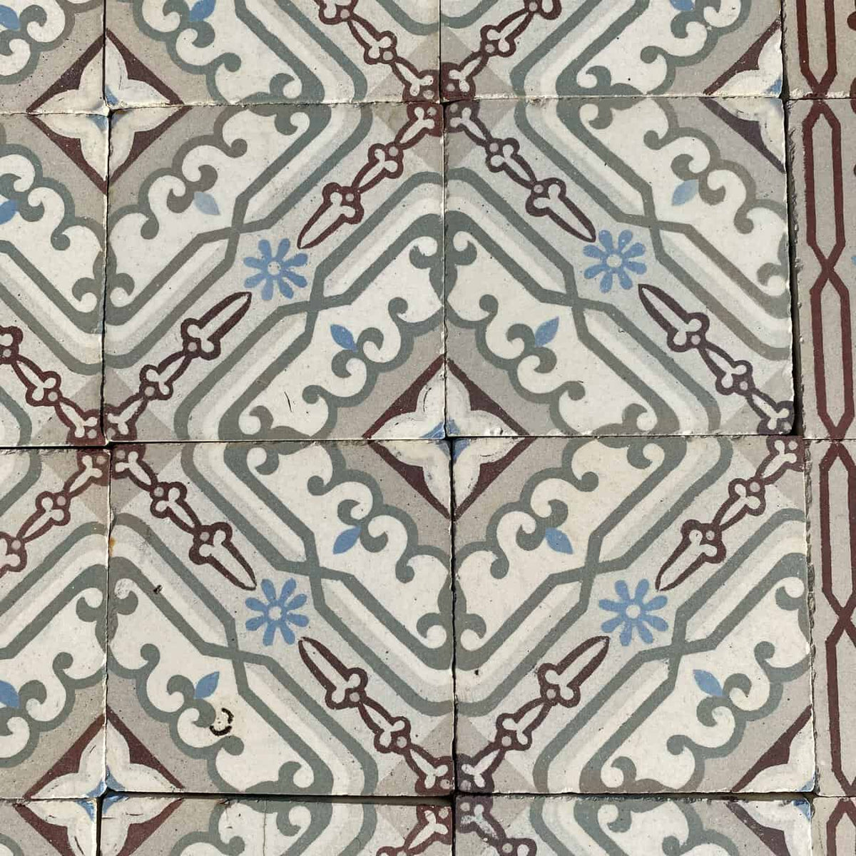Eine Nahaufnahme von Historischen Gründerzeitfliesen mit blassblauen Blumenmustern und kastanienbraunen und grauen geometrischen Formen. Die Fliesen, die jeweils 14 x 14 cm groß sind und etwa 10 Quadratmeter bedecken, bilden ein sich wiederholendes symmetrisches Muster und erzeugen einen Vintage-Dekorationslook, der an historische Fliesen erinnert. Die Fugenlinien sind leicht ungleichmäßig, was den rustikalen Charme der Fliesen noch verstärkt.