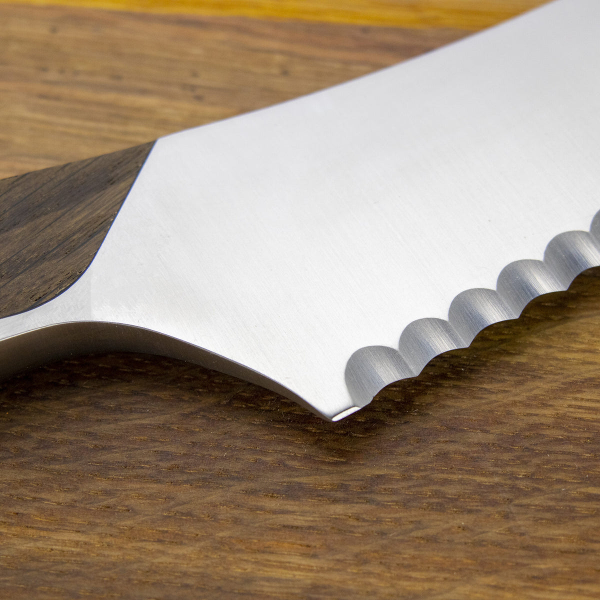 Nahaufnahme eines Güde Synchros Brotmessers mit 32 cm langer Klinge und Holzgriff, ruht auf einem 34 x 21 cm großen Fassholzbrett. Der Fokus liegt auf den Metallverzahnungen, die die scharfen Kanten vor der glatten, polierten Oberfläche der Klinge hervorheben.