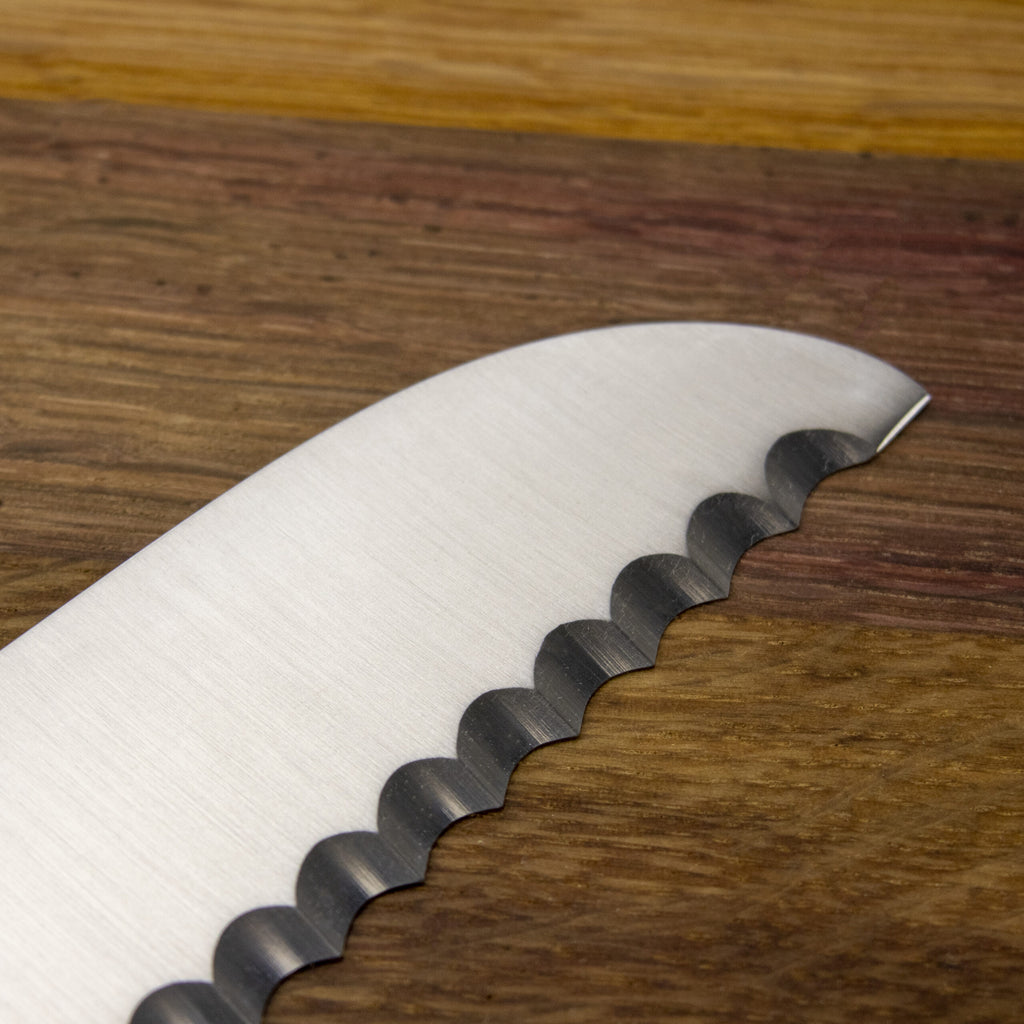 Nahaufnahme eines Güde Synchros Brotmessers mit 32 cm langer Klinge, horizontal auf einem 34 x 21 cm großen Fassholzbrett platziert. Die gewellte Schneide des Messers ist zum Schneiden von zähem oder knusprigem Brot geeignet, und die Holzmaserung des Bretts bietet einen kontrastierenden Hintergrund.