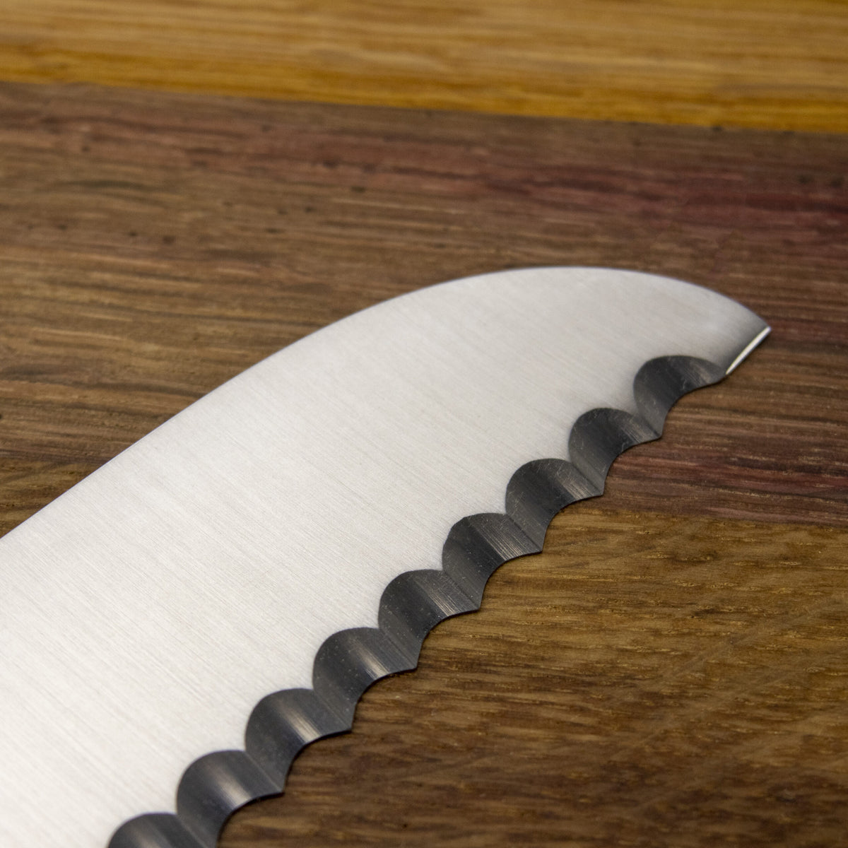 Nahaufnahme eines Güde Synchros Brotmessers mit 32 cm langer Klinge, horizontal auf einem 34 x 21 cm großen Fassholzbrett platziert. Die gewellte Schneide des Messers ist zum Schneiden von zähem oder knusprigem Brot geeignet, und die Holzmaserung des Bretts bietet einen kontrastierenden Hintergrund.