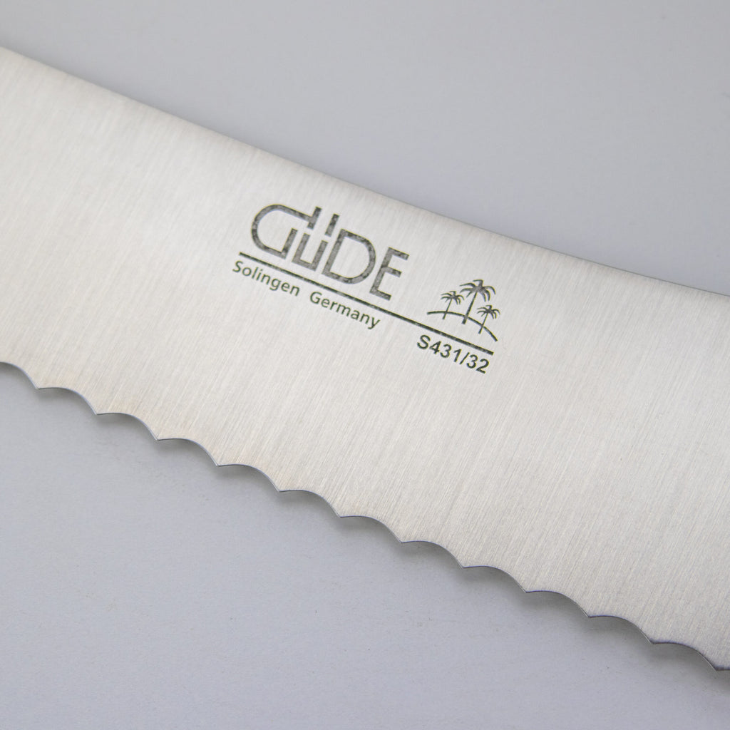 Nahaufnahme einer gezackten Messerklinge mit der Gravur „GÜDE Solingen Germany S431/32“ und einem Bild von zwei Palmen. Das Güde Synchros Brotmesser mit einer 32 cm langen Klinge ruht auf einem hellen, neutralen Hintergrund.