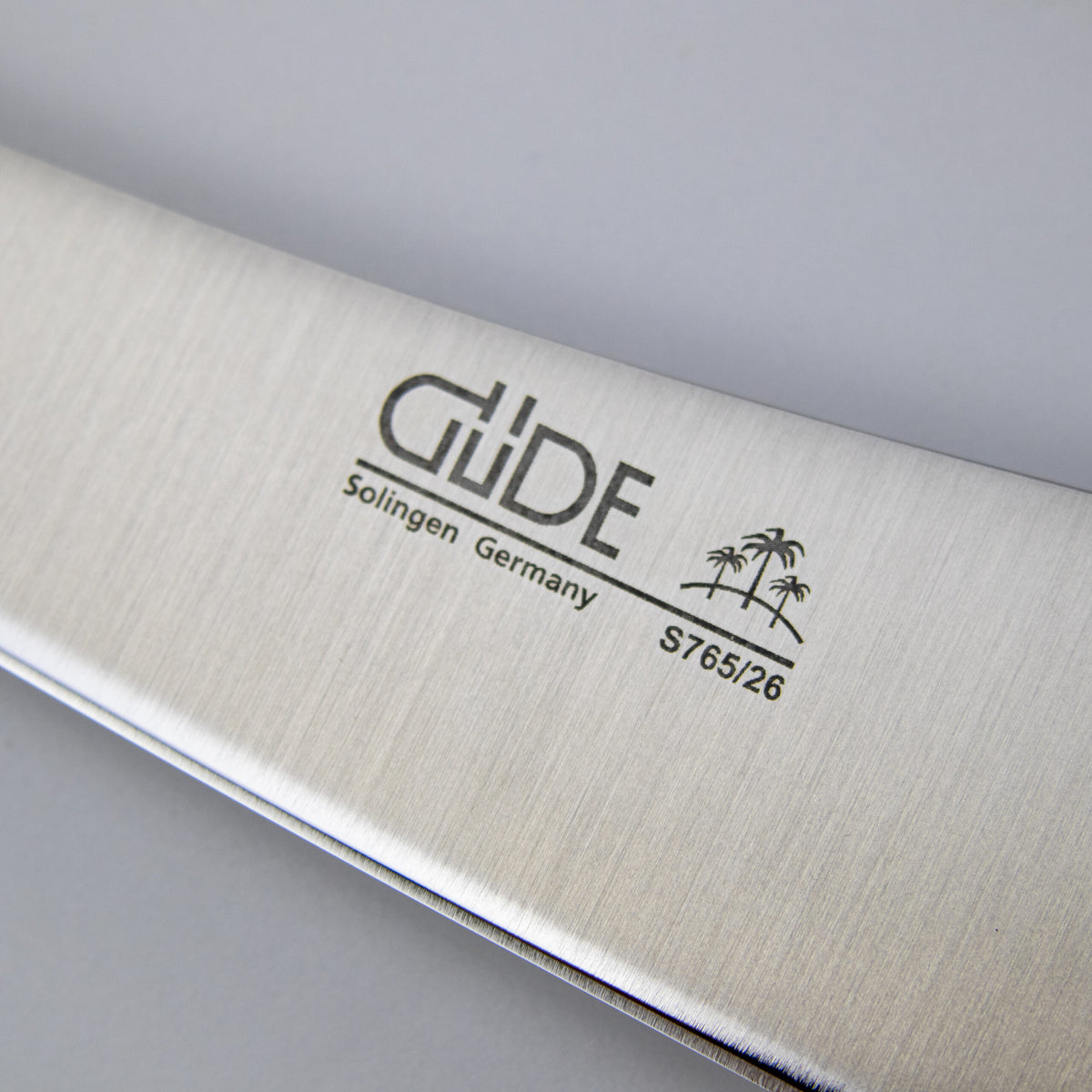 Nahaufnahme der Klinge eines Güde Synchros Tranchiermessers, 26 cm Klinge + Fassholzbrett, mit dem Text „GLiDE Solingen Germany S765/26“ und einer in das Metall eingravierten Grafik von zwei Palmen. Die 26 cm lange Klinge hat eine Oberfläche aus gebürstetem Edelstahl und ist damit ein elegantes und funktionales Werkzeug für jede Küche.