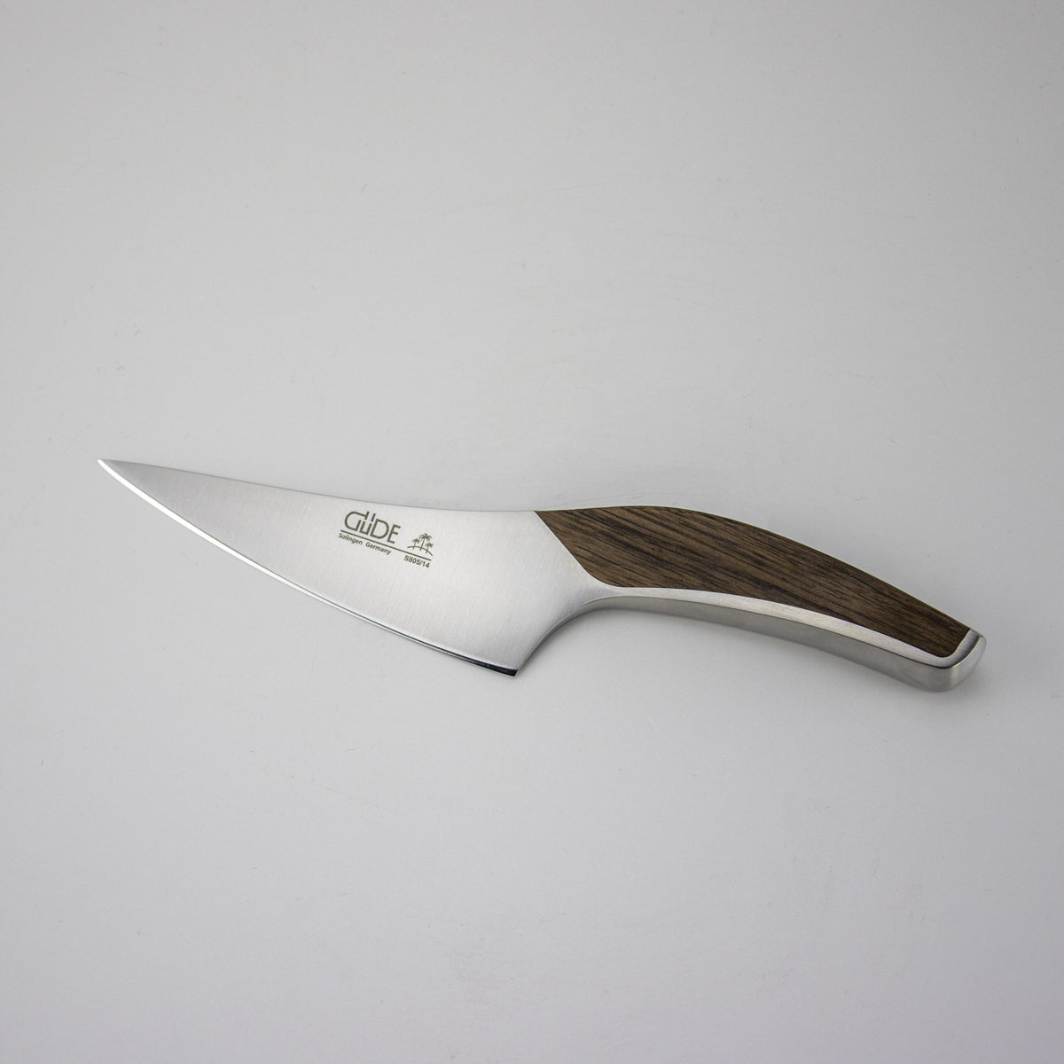 Ein schlichtes Küchenmesser, das Güde Synchros Zubereitungsmesser mit einer 14 cm langen polierten Metallklinge und einem Holzgriff, ruht auf einem schlichten, hellen handgefertigten Fassholzbrett mit den Maßen 34x21 cm. Die Klinge trägt den Markennamen „Güde“ sowie einige zusätzliche kleine Texte und ein Logo.