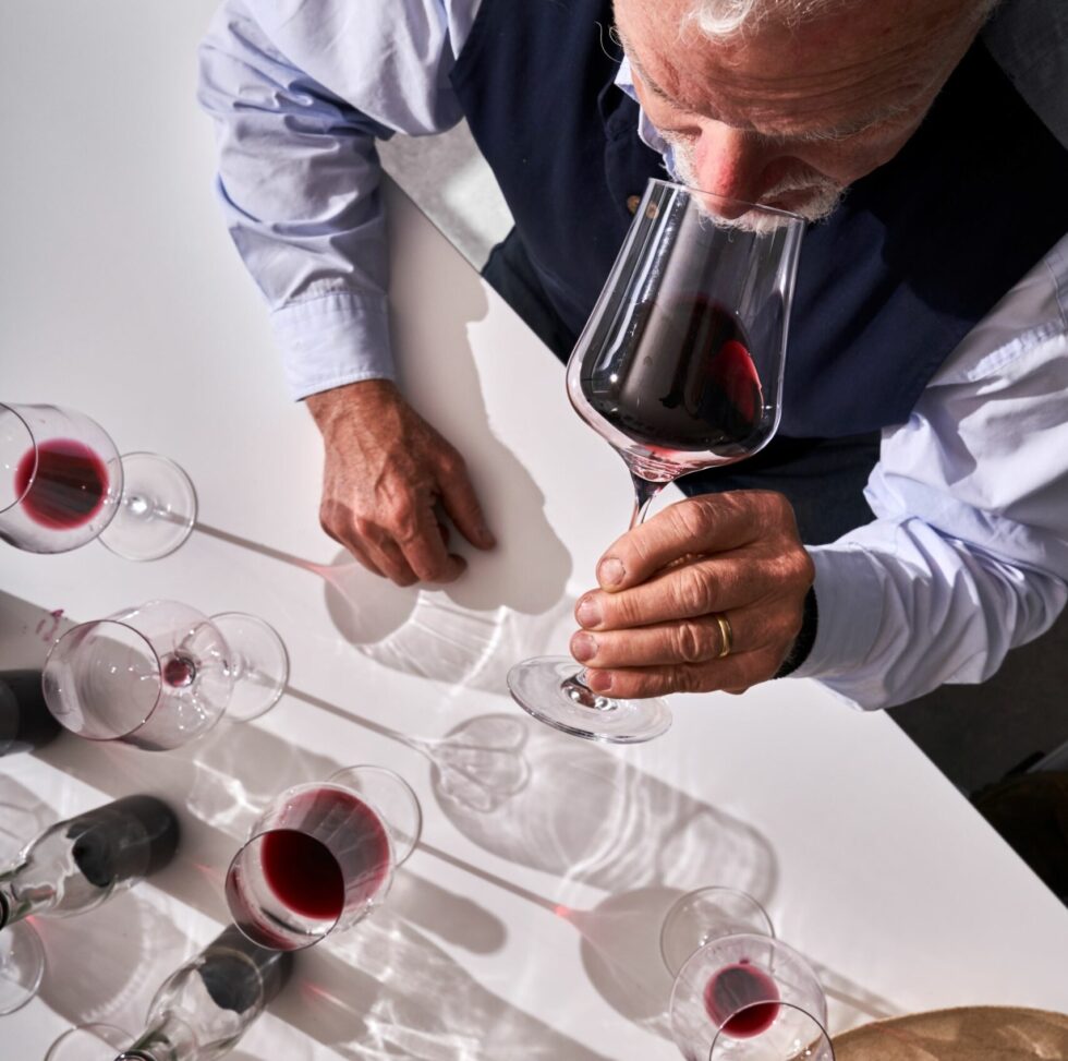 Gabriel Gold, mit seinem auffälligen weißen Haar, nippt nachdenklich an einem Gabriel Gold Edition Weinglas Universalglas 90 Gramm, mundgeblasen und spülmaschinenfest, während er am Tisch sitzt. Mehrere andere Gläser Rotwein stehen in der Nähe, ihre Schatten und Spiegelbilder tanzen unter dem Deckenlicht.