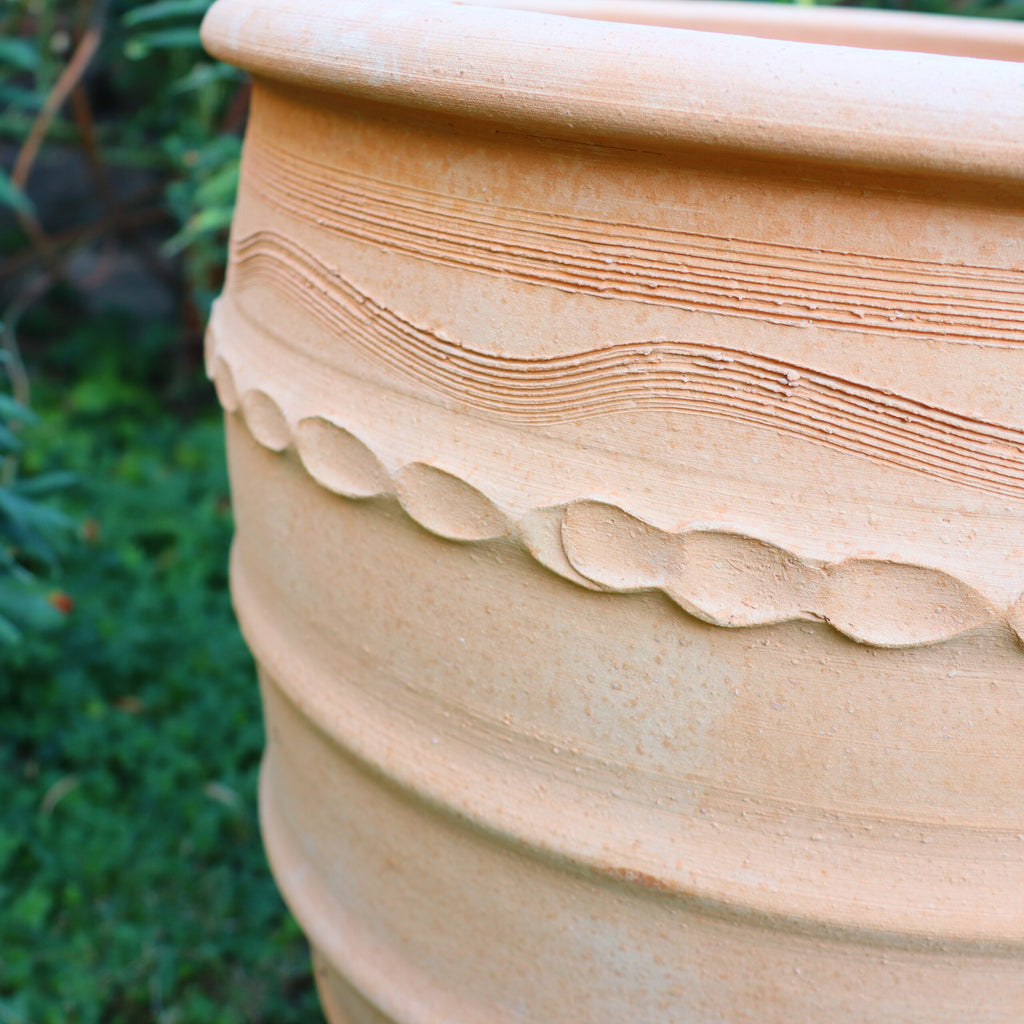Eine Nahaufnahme des Terrakotta-Blumentopfs „Granatum“, einem großen, beigen 50-cm-Topf mit komplizierten, strukturierten Mustern mit Wellenlinien und kreisförmigen Motiven. Der Topf steht im Freien vor einem verschwommenen grünen Laubhintergrund.