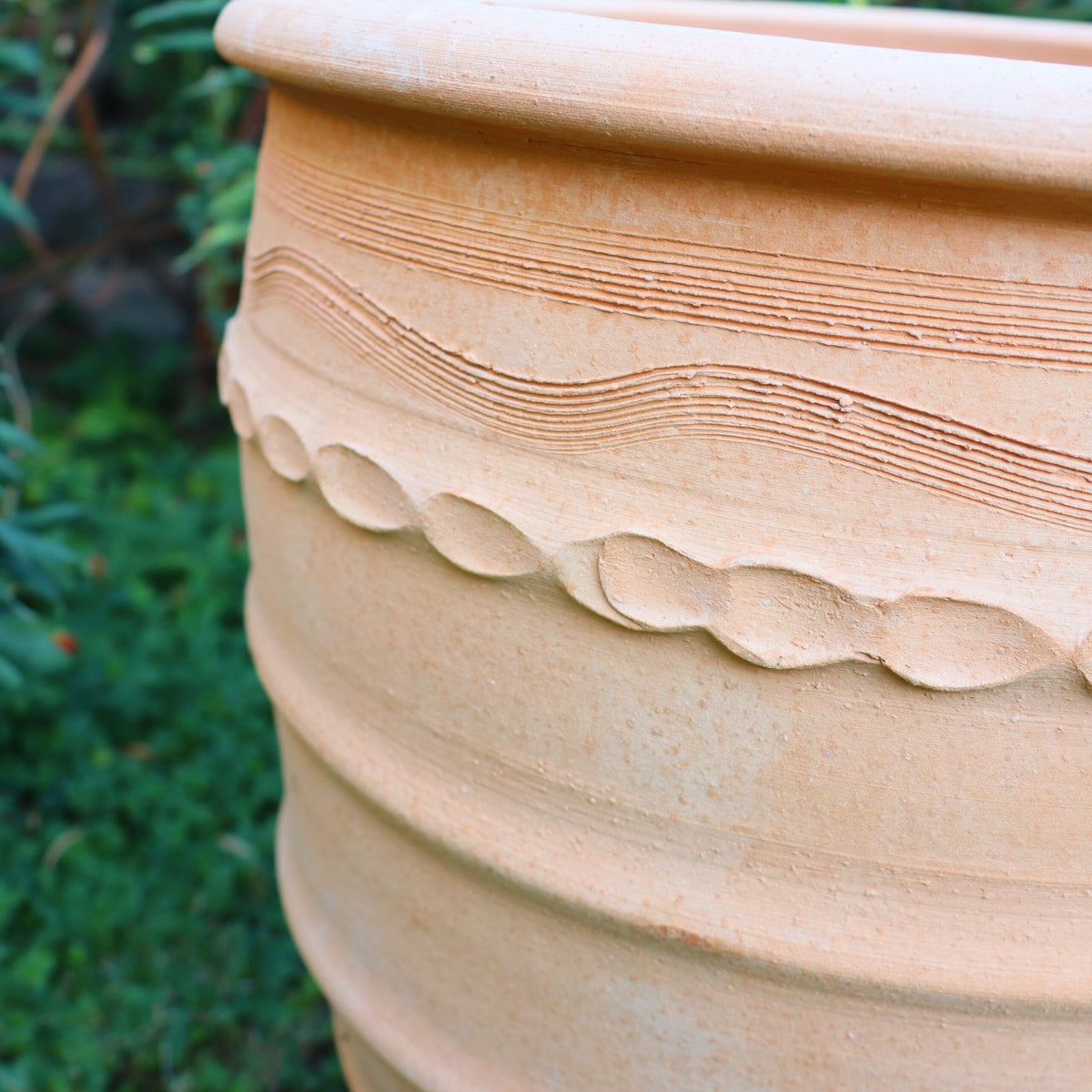 Eine Nahaufnahme des Terrakotta-Blumentopfs „Granatum“, einem großen, beigen 50-cm-Topf mit komplizierten, strukturierten Mustern mit Wellenlinien und kreisförmigen Motiven. Der Topf steht im Freien vor einem verschwommenen grünen Laubhintergrund.