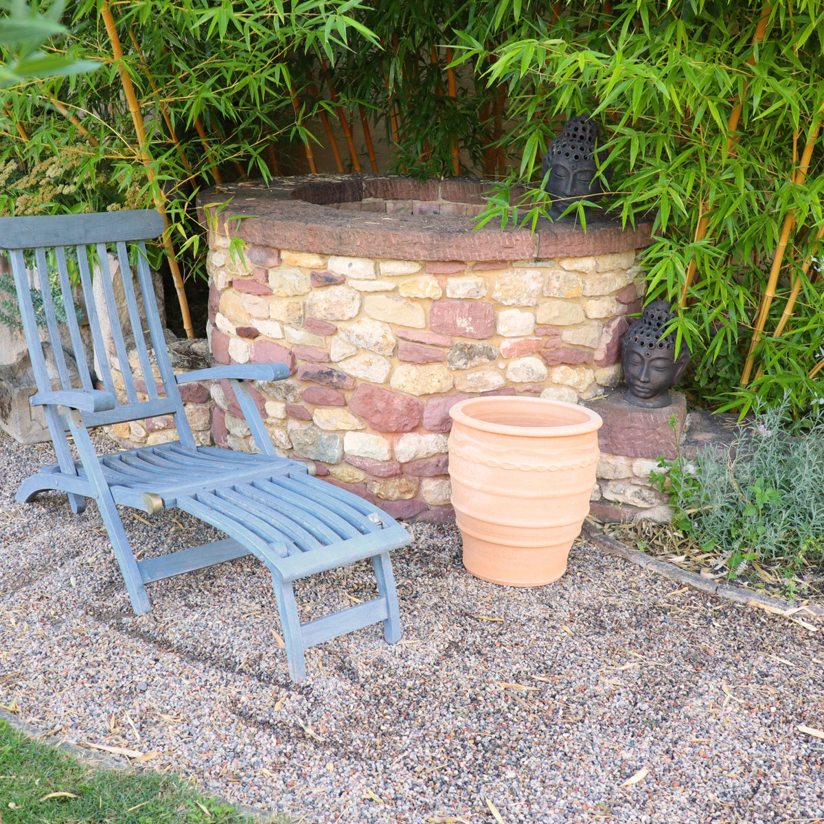 Eine Gartenszene mit einem blauen Liegestuhl aus Holz auf einer Kiesterrasse neben einem Steinbrunnen. Ein großer Blumentopf aus Terrakotta „Granatum“ mit 50 cm Durchmesser steht neben dem Brunnen, während Bambuspflanzen und zwei Buddha-Kopfstatuen einen ruhigen Hintergrund bilden.