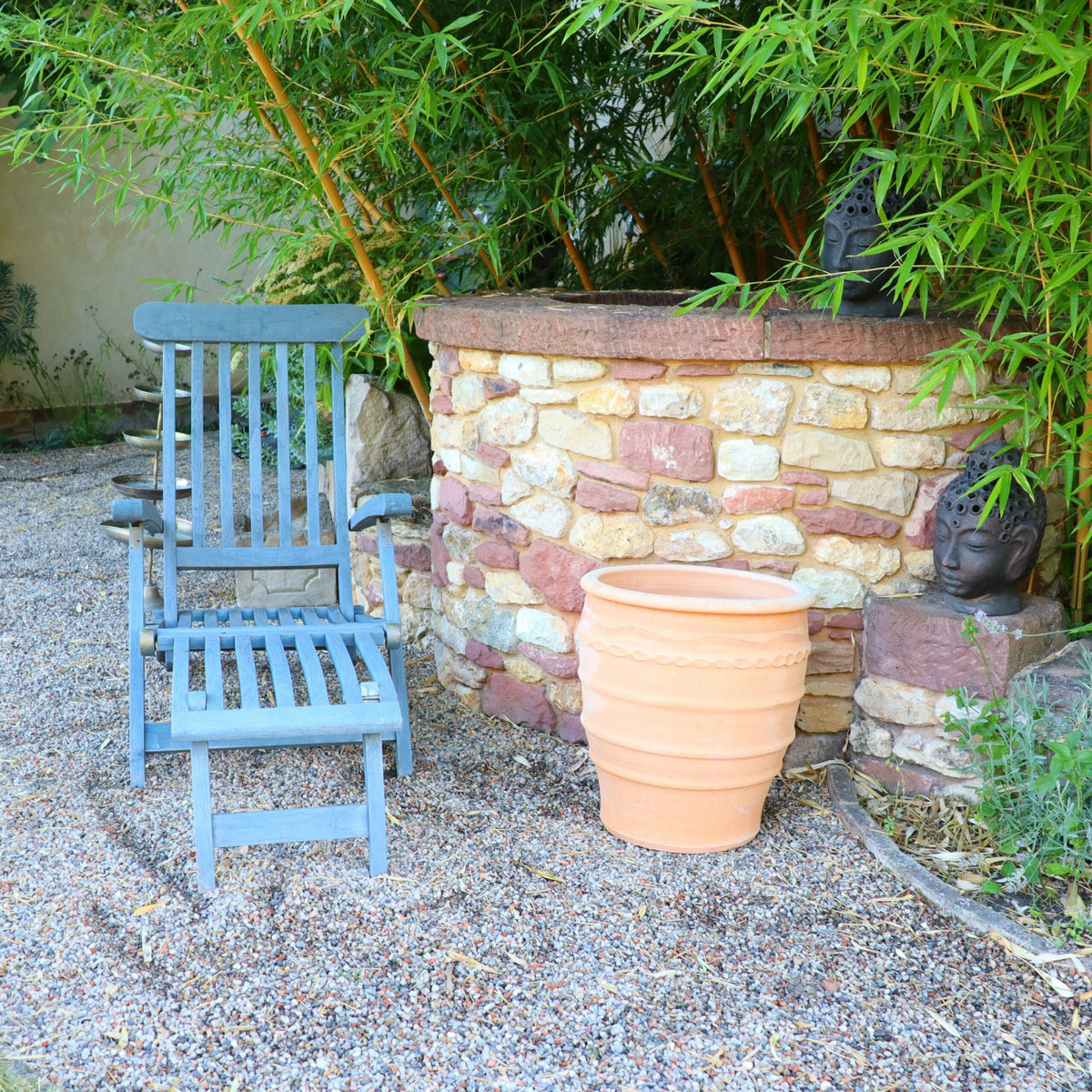 Eine Szene im Freien mit einem blauen Holzstuhl auf Kies neben einem 50 cm hohen Blumentopf aus Terrakotta „Granatum“ und einer niedrigen Steinmauer. Hinter der Mauer wachsen grüne Bambuspflanzen und neben dem Blumentopf ist teilweise eine kleine Buddhastatue zu sehen.
