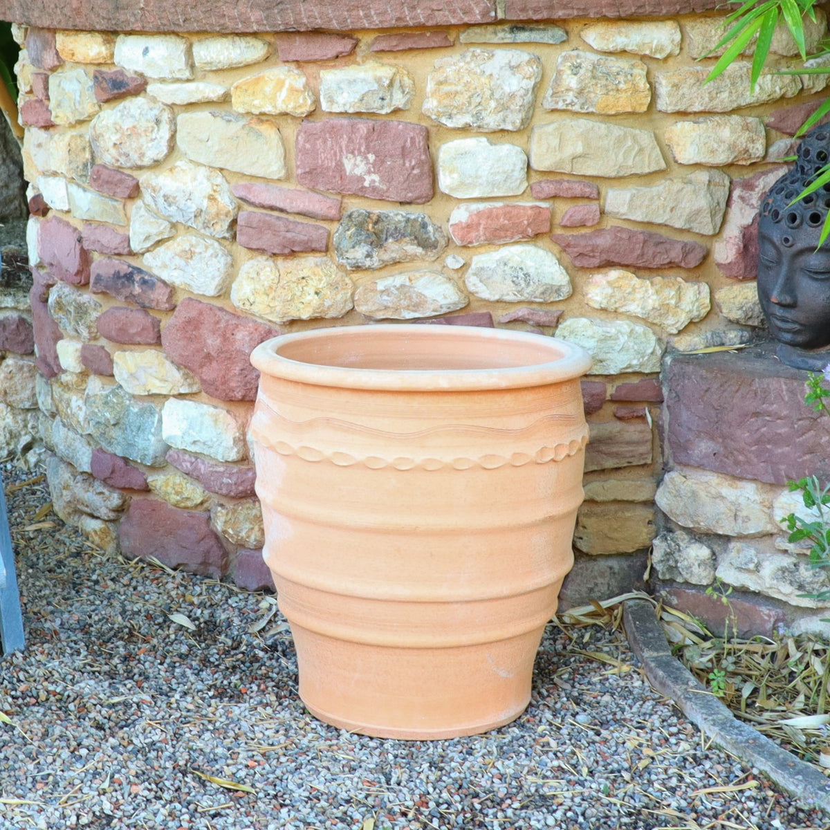 Ein großer, leerer Terrakotta Blumentopf „Granatum“, 50cm steht auf einer Kiesfläche vor einer rustikalen Steinmauer. Rechts, teilweise sichtbar, steht eine dekorative Buddhakopf-Skulptur inmitten üppigen Grüns.