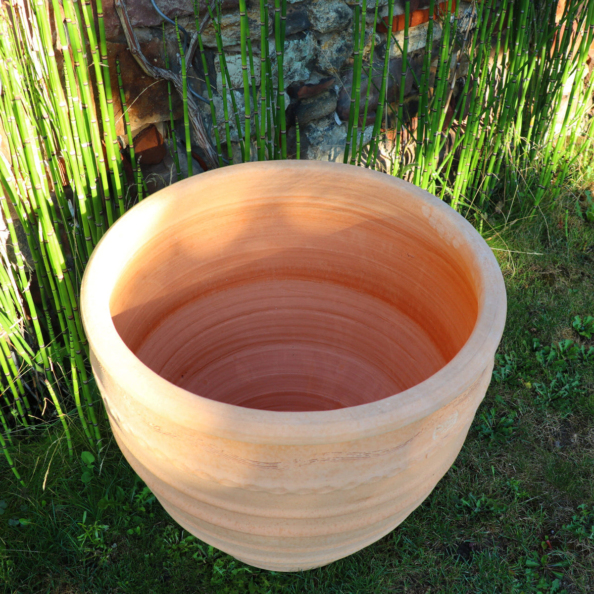Ein großer, leerer frostfester Blumentopf „Granatum“ 60 cm steht auf einem Grasfleck in der Nähe einer Steinmauer und Bambusstäben. Der Blumentopf hat eine glatte, runde Form mit dezenten horizontalen Rillen. Das Sonnenlicht wirft weiche Schatten und hebt die Textur des Granatums hervor.