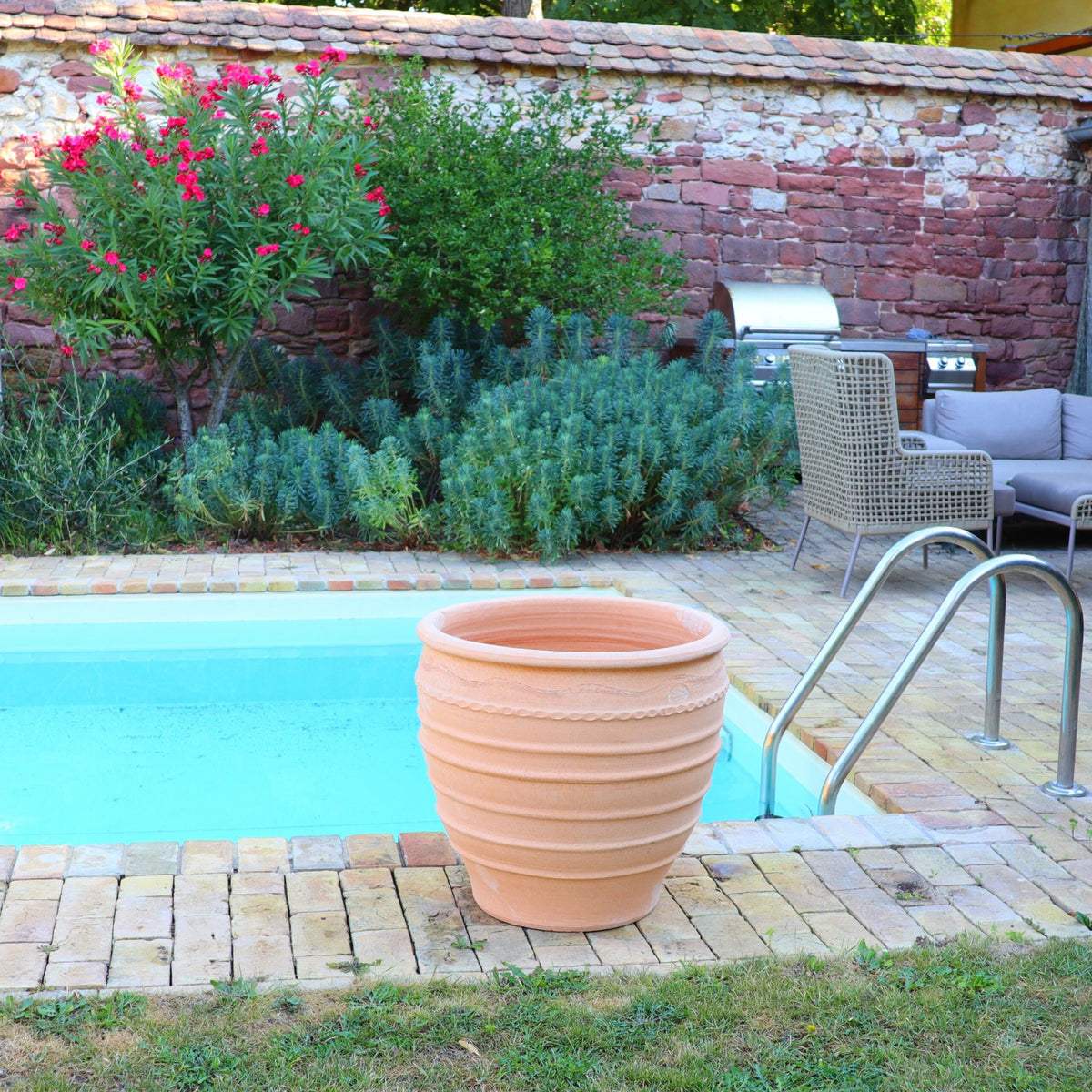 Ein ruhiger Hinterhof mit einem kleinen, einladenden Swimmingpool, umgeben von beigen Ziegeln. Neben dem Pool steht ein großer frostfester Blumentopf „Granatum“ (60 cm) aus Terrakotta. Üppiges Grün, darunter hohe Büsche und blühende Pflanzen, bereichert die ruhige Umgebung. Im Hintergrund stehen Korbmöbel und ein Grill.