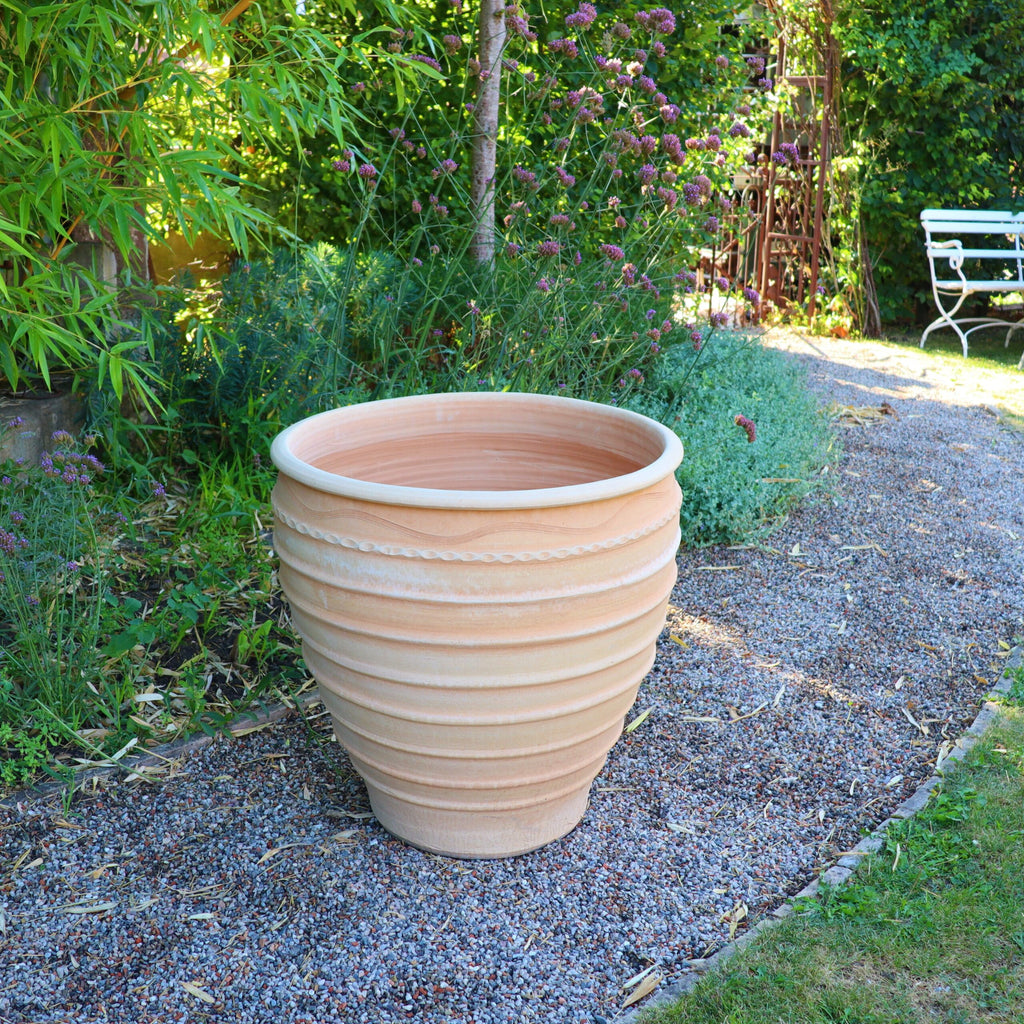 Ein großer Winterharter Pflanzkübel gross „Granatum“, 70 cm, mit gerippter Verzierung steht auf einem Kiesweg in einem Garten. Um den Topf herum sind verschiedene Pflanzen und Grünpflanzen, im Hintergrund ist teilweise eine weiße Gartenbank inmitten üppigen Laubes zu sehen.
