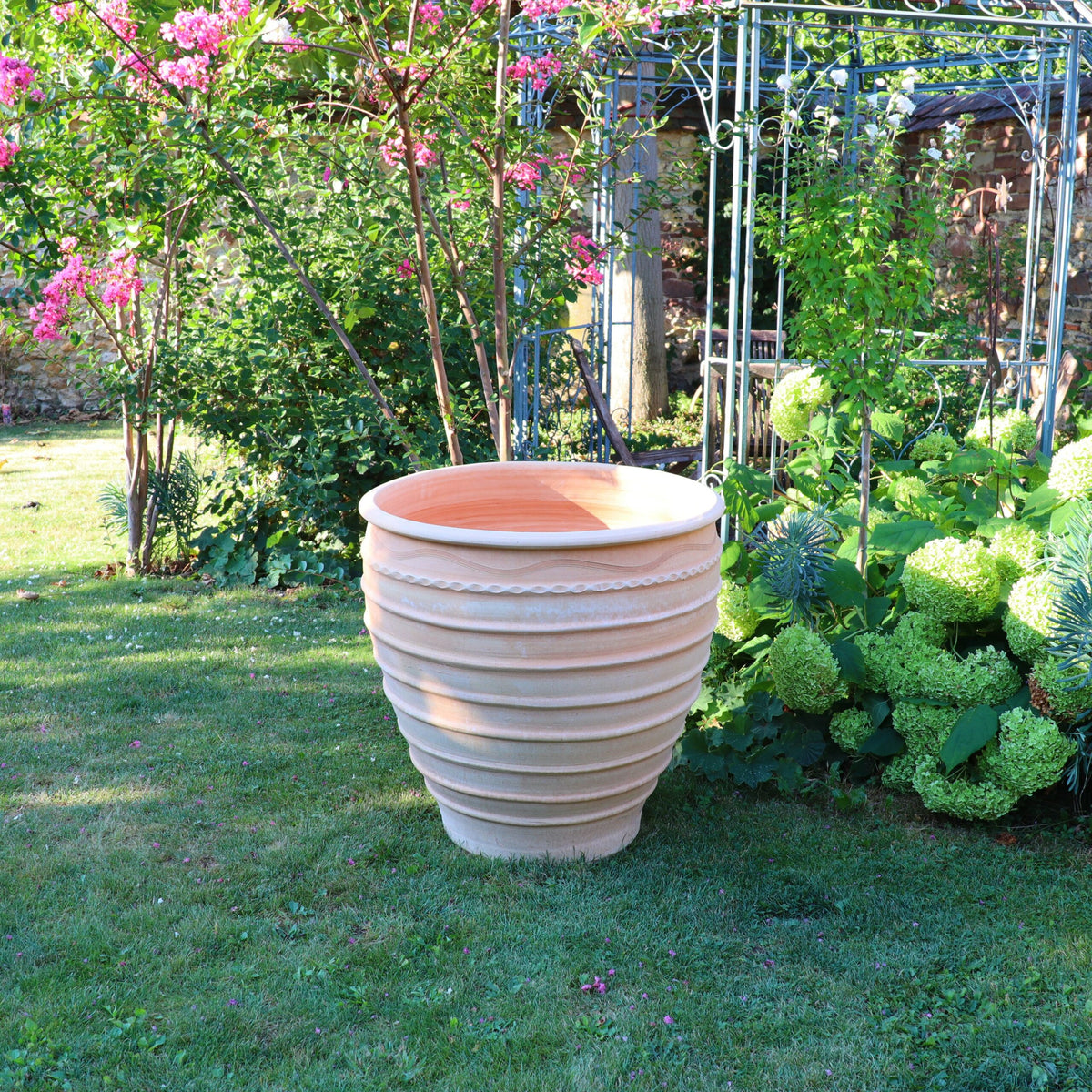 Ein großer, beigefarbener Winterharter Pflanzkübel „Granatum“, 70 cm, mit horizontalen Rillen steht auf einem gepflegten Rasen. Der Hintergrund zeigt üppiges Grün, blühende rosa Granatum-Blüten und eine mit Kletterpflanzen bedeckte Metallgitterstruktur. Die Szene scheint Teil eines ruhigen Gartens zu sein.
