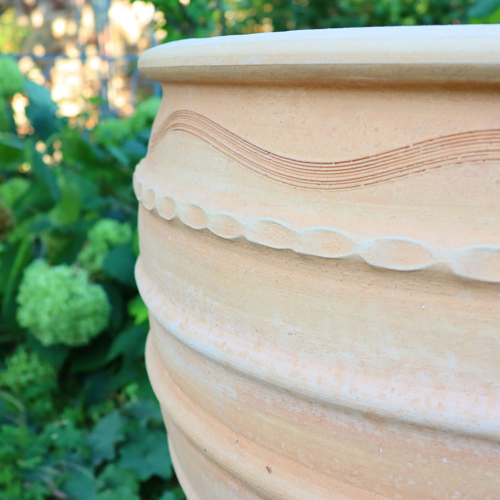 Nahaufnahme eines strukturierten, beigen Terrakotta-Topfes mit komplizierten Mustern, platziert in einer Gartenumgebung. Der Hintergrund zeigt grünes Laub und Büschel hellgrüner Blüten, teilweise unscharf, um die Details des Winterharter Pflanzkübels gross „Granatum“, 70 cm, hervorzuheben.