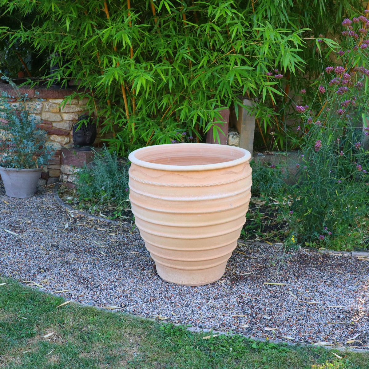 Ein großer, leerer gerippter Terrakotta-Pflanzkübel „Granatum“ (70 cm) von Winterhart steht auf einem Kiesweg in einem üppigen Garten. Der Topf ist von verschiedenen Grünpflanzen umgeben, darunter hoher Bambus und violette Blumen. Im Hintergrund ist eine Steinmauer zu sehen.
