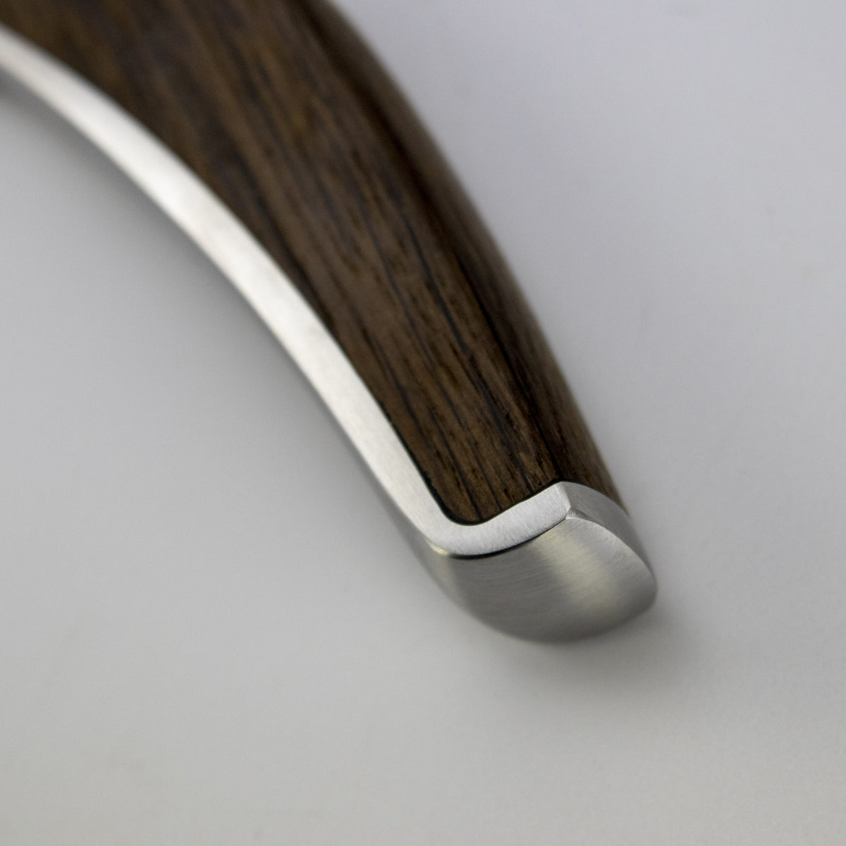 Nahaufnahme des Griffs eines Güde Synchros Brotmessers, 32 cm Klinge + 34x21 cm Fassholzbrett, zeigt den fließenden Übergang von poliertem Metall zu einem dunklen Holzfinish. Die Klinge erscheint glatt und glänzend, während das Holzsegment eine braune, strukturierte Maserung aufweist. Der Hintergrund hat eine neutrale, helle Farbe.