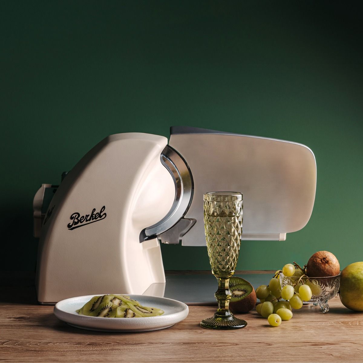 A Berkel Home Line 200 Plus creme MATT Aufschnittmaschine + handgefertigtes Schneidebrett Unikat steht auf einem Holztisch mit Fruchtarrangement und grünem Hintergrund.