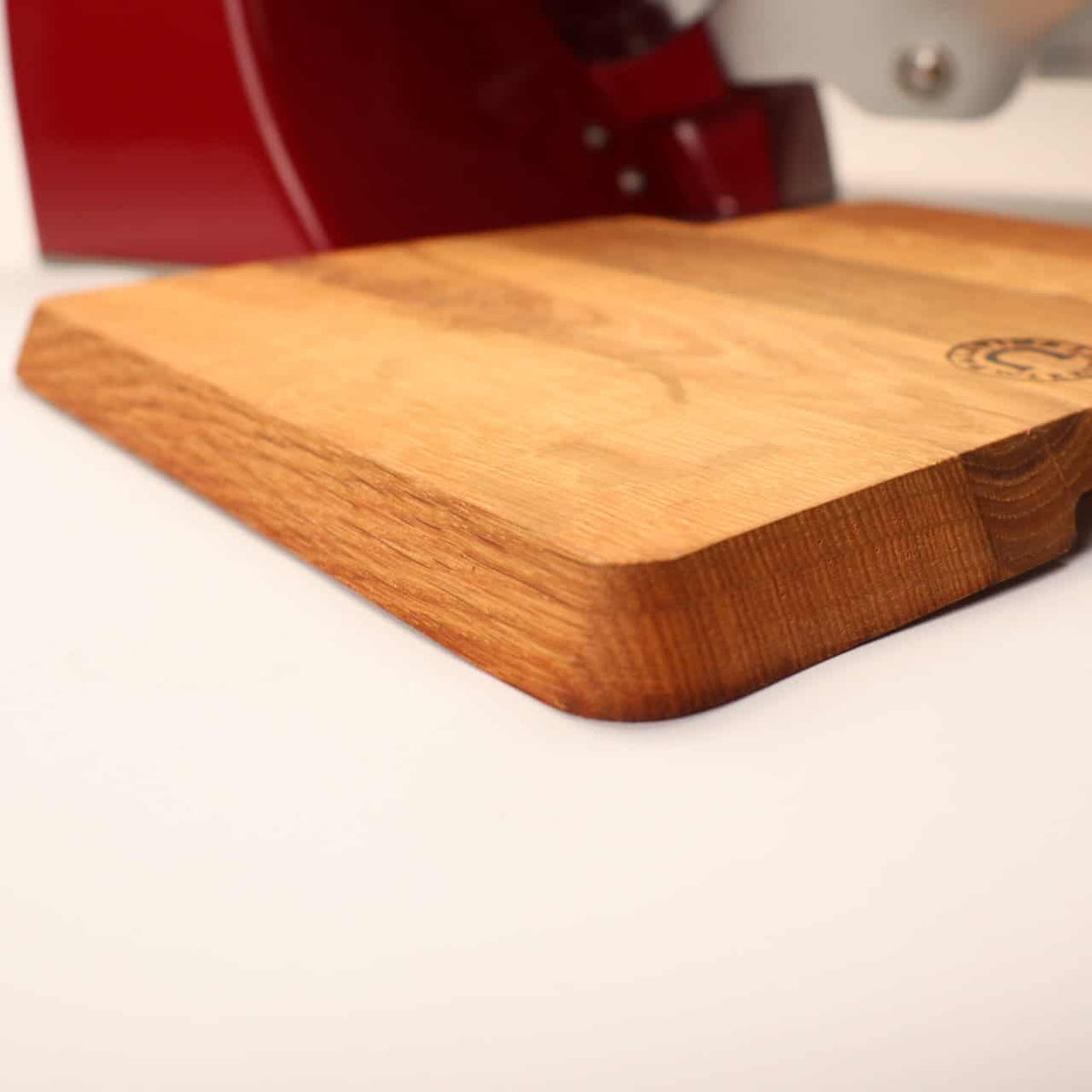 Nahaufnahme des Palatina-Werkstatt Einlegebretts passend für Berkel Home Line 250 auf hellem Untergrund. Das Holzschneidebrett hat eine glatte Oberfläche mit abgerundeten Kanten und einem eingeprägten Logo der Palatina-Werkstatt in einer Ecke. Im Hintergrund ist ein Teil einer roten Berkel Home Line 250-Maschine teilweise sichtbar.