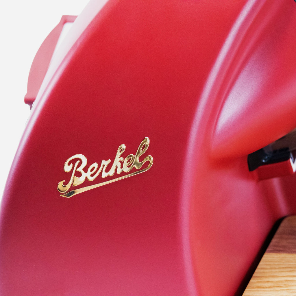 Nahaufnahme der Berkel Home Line 200 Plus rot MATT Aufschnittmaschine - neues Modell - mit goldenem Logo auf der gewölbten roten Oberfläche und einem Teil des einzigartigen handgefertigten Schneidebretts.
