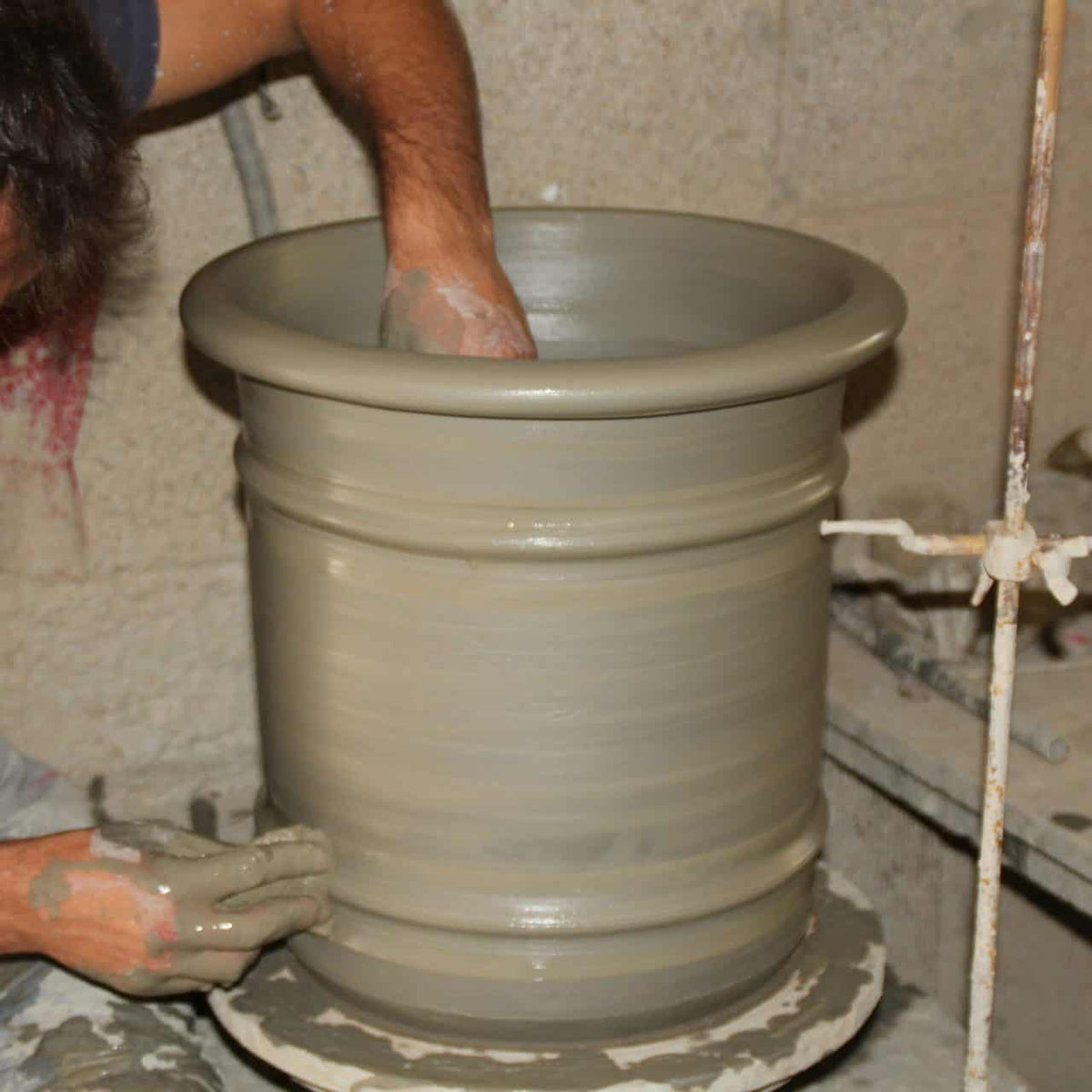 Eine Person formt einen großen Übertopf aus Terrakotta „Canna“ (Höhe 50 cm) auf einer Töpferscheibe und glättet und formt mit den Händen die Seiten des Topfes. Der Arm des Töpfers ist teilweise sichtbar, da er in einer Werkstatt, die wie eine Töpferwerkstatt aussieht, konzentriert arbeitet.