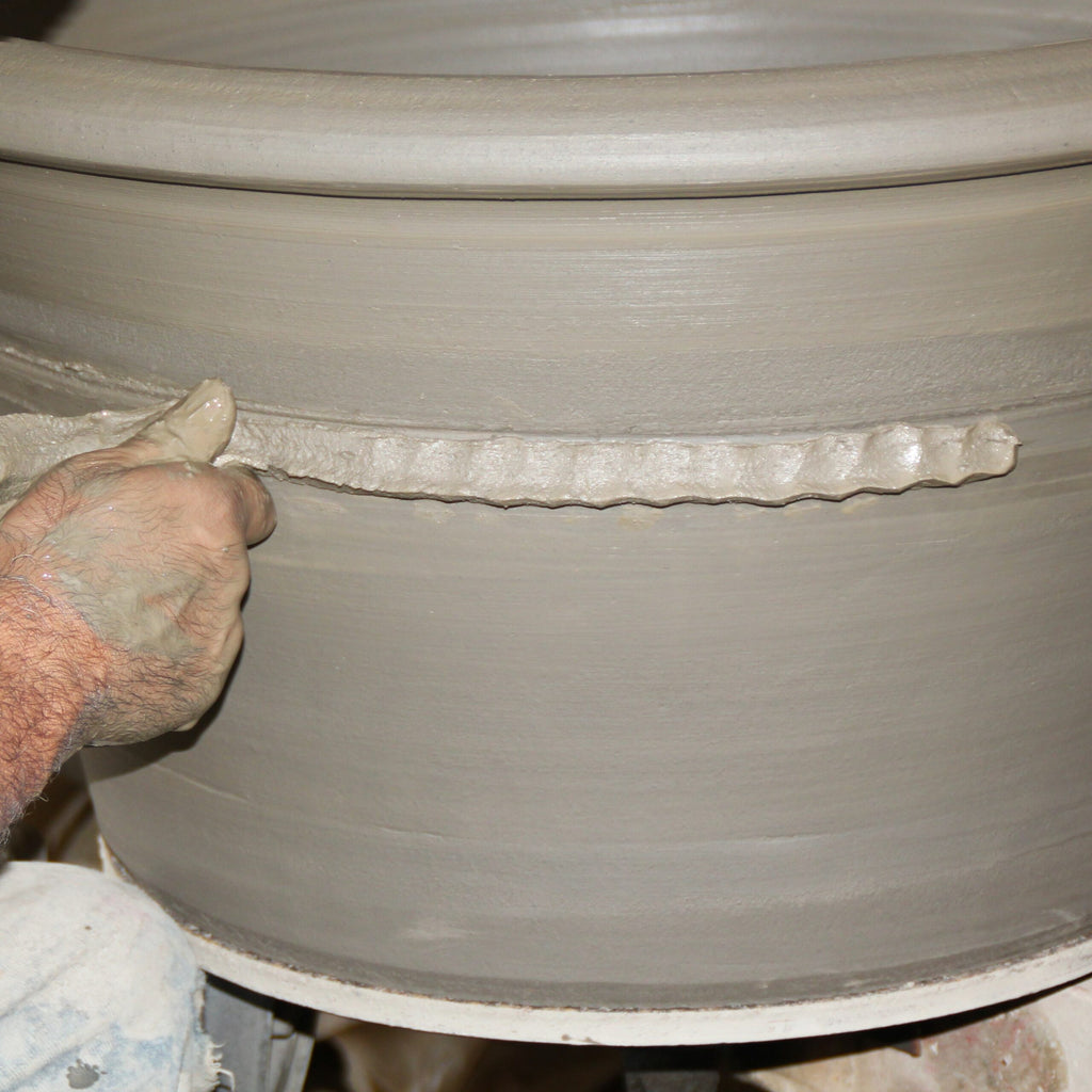 Eine Nahaufnahme zeigt die Hände einer Person, die einen Streifen feuchten Tons auf einen großen, glatten Terrakotta-Pflanzkübel „Acanthus“, H=50 cm, formt. Hände und Ton sind mit Schlamm bedeckt, und der Pflanzkübel befindet sich noch auf einer Töpferscheibe. Die Szene fängt den detaillierten und taktilen Prozess der Herstellung von Tontöpferei ein.