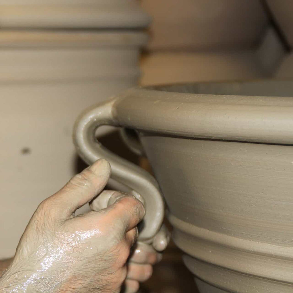 Nahaufnahme der Hände, die einen Henkel an einem Pflanzkübel "Salvia 2" (H=25 cm) aus nassem Ton formen, wobei das handwerkliche Können und der keramische Herstellungsprozess auf einer Töpferscheibe hervorgehoben werden.