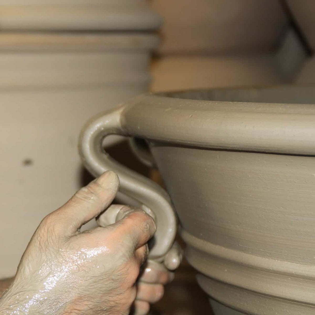 Nahaufnahme der Hände, die einen Henkel an einem Pflanzkübel "Salvia 2" (H=25 cm) aus nassem Ton formen, wobei das handwerkliche Können und der keramische Herstellungsprozess auf einer Töpferscheibe hervorgehoben werden.