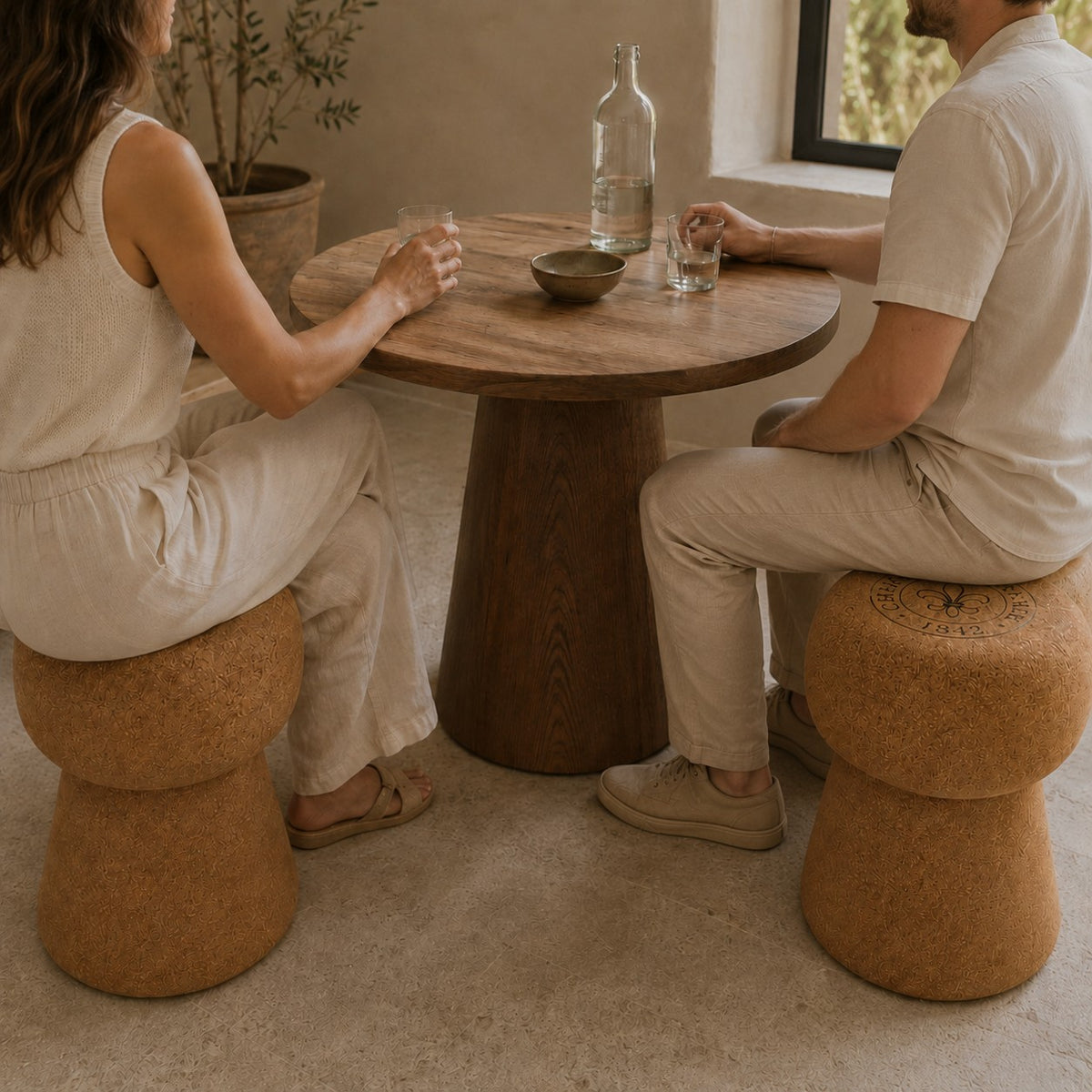 Zwei Personen in heller, neutraler Kleidung sitzen auf einem hochwertigen Hocker aus Kork in Form eines Champagnerkorkens an einem runden Holztisch mit Wassergläsern, einer Glasflasche und einer Schale. Die minimalistische Umgebung ist natürlich beleuchtet.