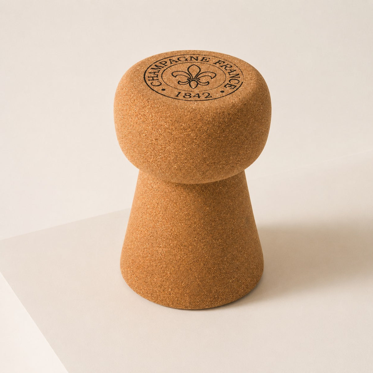 Ein hochwertiger Hocker aus Kork in Form eines Champagnerkorkens steht aufrecht auf einer hellen Fläche. Der Deckel zeigt die Prägung "CHAMPAGNE FRANCE 1842" und ein Fleur-de-Lis Motiv.