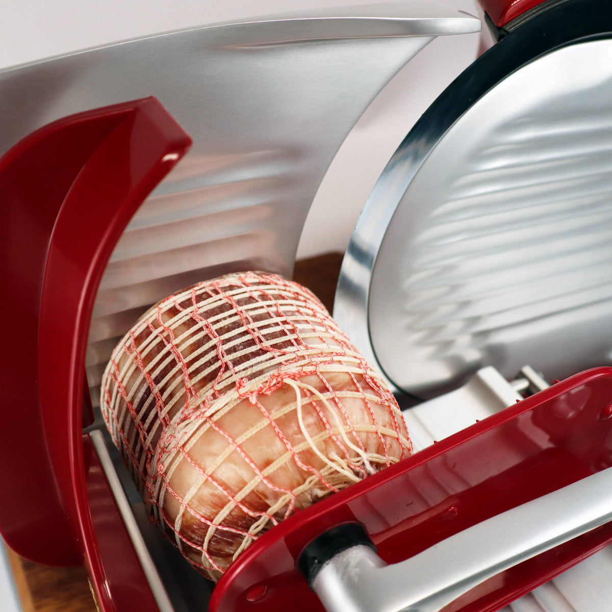 Eine Nahaufnahme der Berkel Home Line 250 Plus rot Aufschnittmaschine (neues Modell) mit Wellenschliff-Bausatz für Brot und einem handgefertigten einzigartigen Schneidebrett, bereit zum Schneiden von in Netz eingewickeltem Wurstwarenfleisch. Mit stilvollen roten und silbernen Teilen für präzises Schneiden.