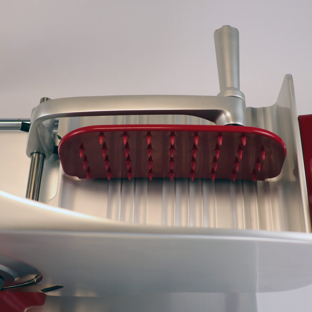 Nahaufnahme eines metallischen und roten Fleischklopfers über einem gerillten Tablett, mit einer unscharfen Berkel Home Line 200 Plus rot Aufschnittmaschine mit handgefertigtem Unikat-Schneidebrett & Wellenschliff-Bausatz für Brot im Hintergrund.