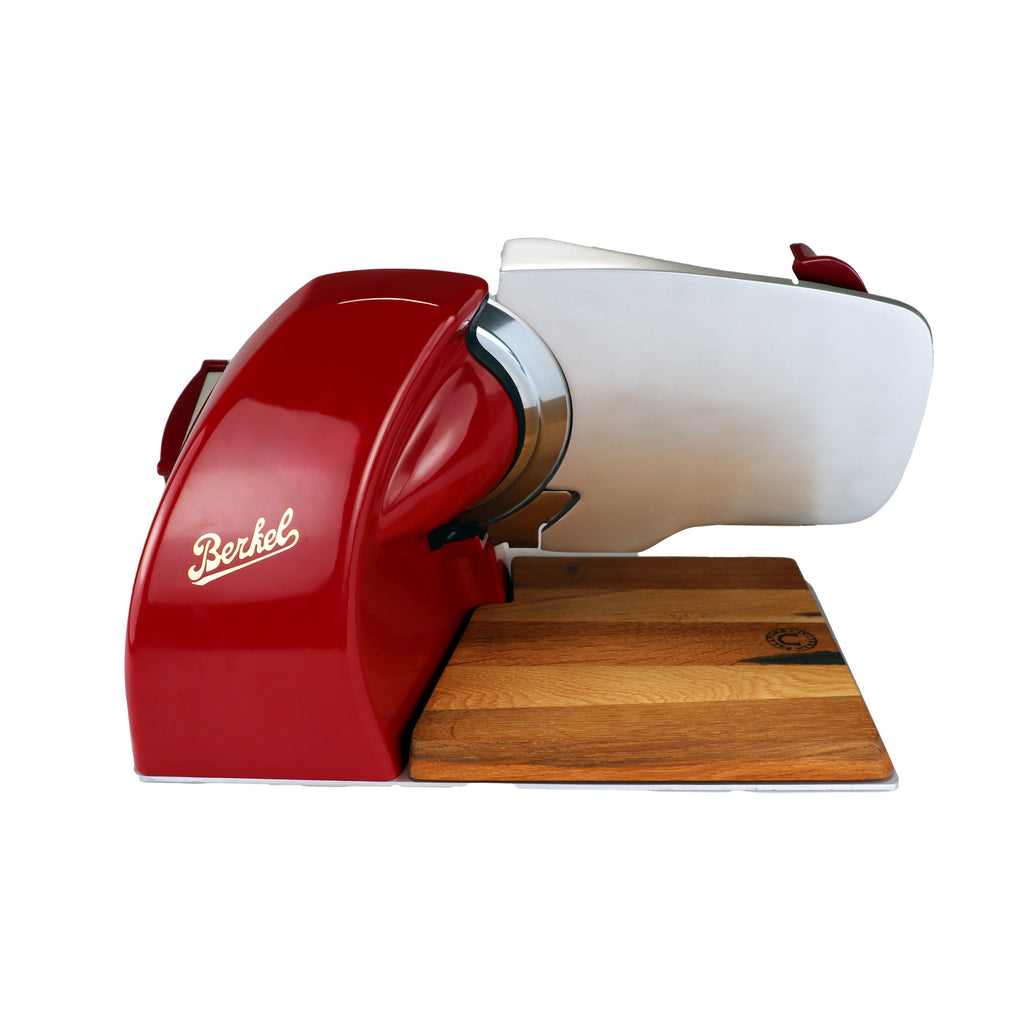 Die Berkel Home Line 200 Plus rot - Aufschnittmaschine neues Modell mit handgefertigtem Unikat-Schneidebrett & Wellenschliff-Bausatz für Brot zeichnet sich durch eine glänzende Klinge und ein hölzernes Schneidebrett aus, das von der Seite auf weißem Hintergrund gezeigt wird.