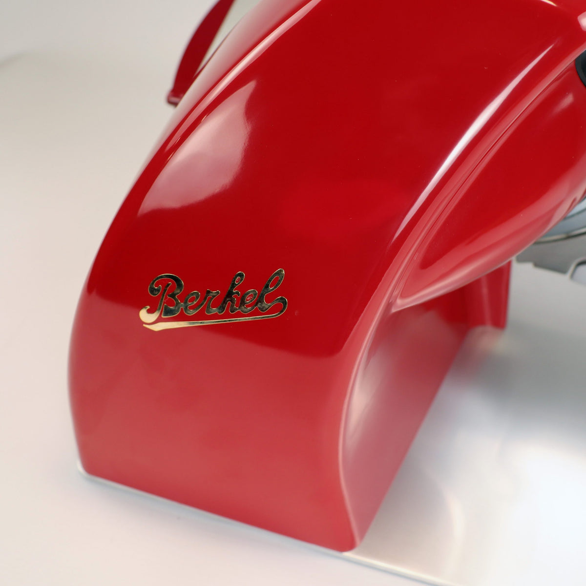 Eine Nahaufnahme der glänzend roten Berkel Home Line 200 Plus Aufschnittmaschine, neues Modell, mit goldfarbenem Logo, handgefertigtem Unikat-Schneidebrett und Wellenschliff-Bausatz für Brot vor weißem Hintergrund.