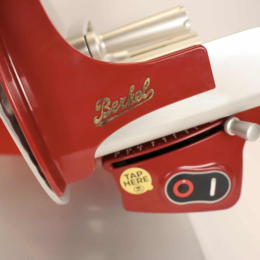 Nahaufnahme einer Berkel Home Line 200 Plus rot, die das metallische „Berkel“-Logo in eleganter Schrift hervorhebt. Unter dem Logo befindet sich ein Drehknopf mit einer „Hier tippen“-Taste, die anzeigt, wo der Schneidemechanismus bedient oder eingestellt werden kann. Diese neue Modell-Aufschnittmaschine wird mit einem 100 % handgefertigten Schneidebrett Unikat für verbesserte Funktionalität und Stil geliefert.