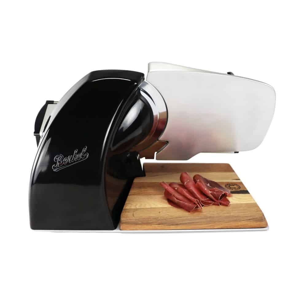 Eine Berkel Home Line 200 Plus Schwarz, Aufschnittmaschine - Neues Modell - Schneidemaschine steht auf einem 100 % handgefertigten Schneidebrett Unikat. Mehrere dünne Wurstscheiben liegen ordentlich auf dem Brett vor der Aufschnittmaschine. Die Aufschnittmaschine hat ein schlankes und modernes Design mit einer teilweise sichtbaren glänzenden Klinge.