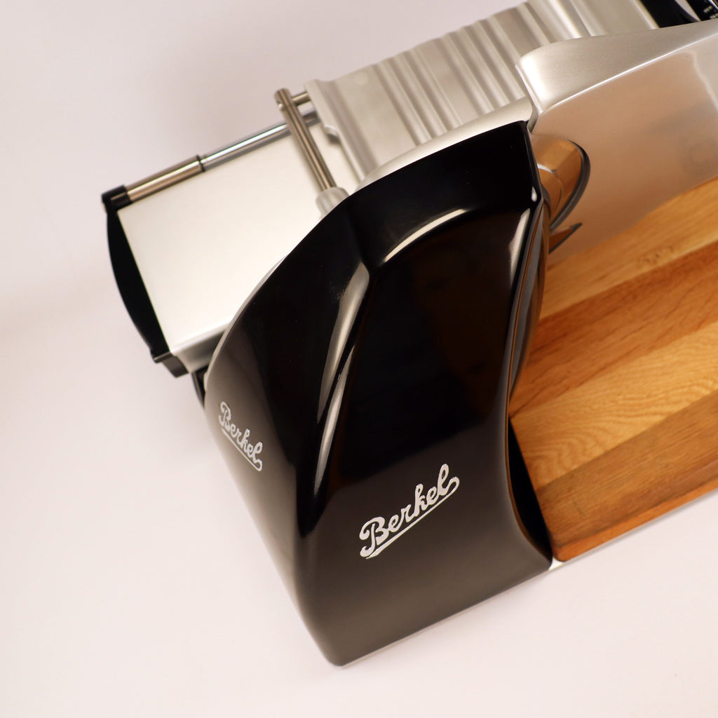 Eine Draufsicht auf die Berkel Home Line 200 Plus schwarz Aufschnittmaschine, das neue Modell mit einem einzigartigen handgefertigten Schneidebrett und einem Brot-Wellenschneide-Set, das sich durch ein elegantes schwarz-silbernes Design auf weißem Hintergrund auszeichnet.